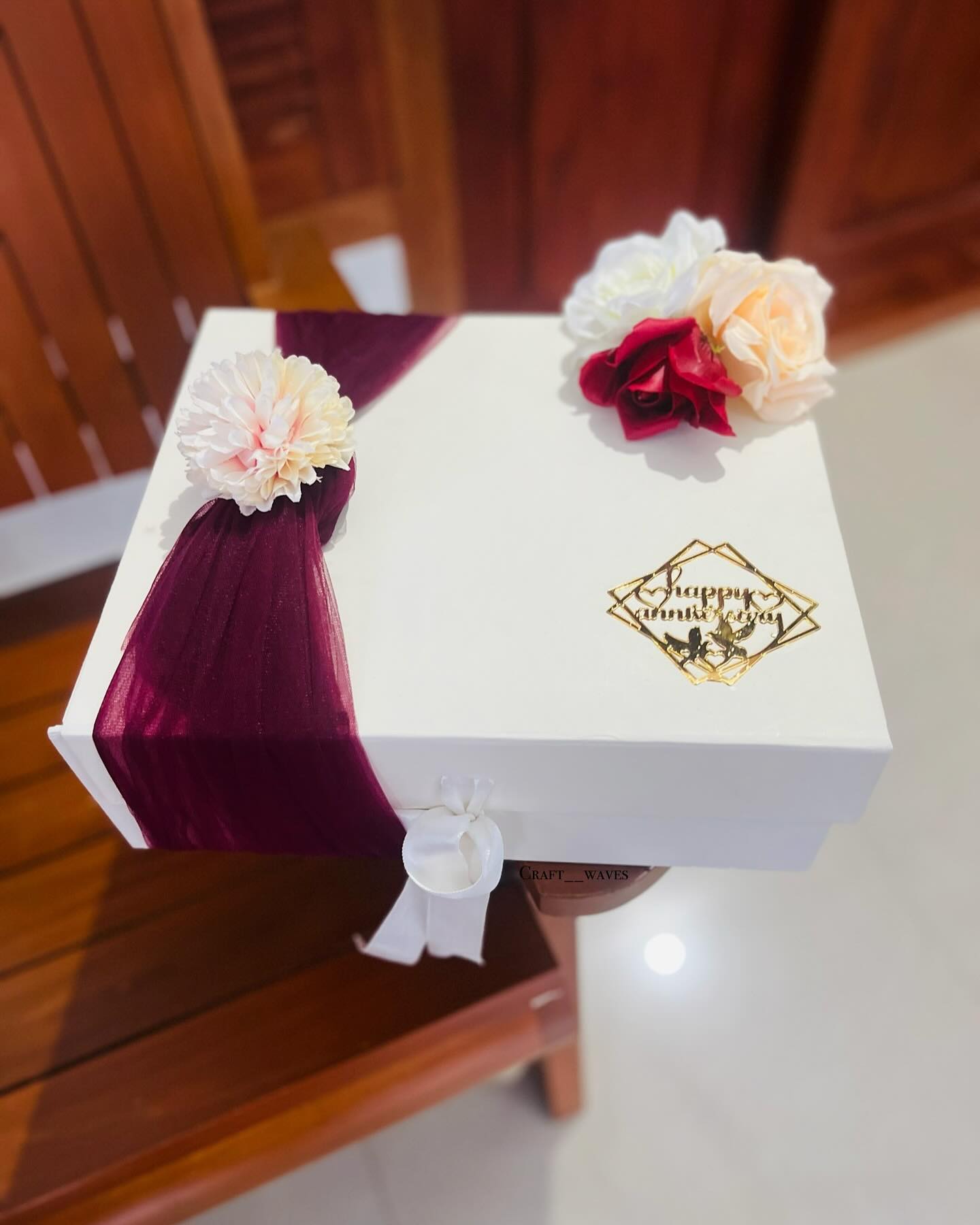 A white keepsake box wrapped in a burgundy tulle bow with cream and red blooms and a gold 'Happy Anniversary' charm.