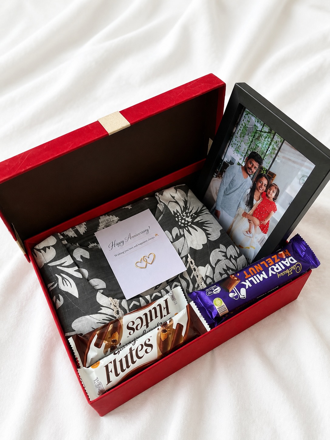 A red anniversary hamper with a printed kurta set, a family photo frame, and assorted chocolates.