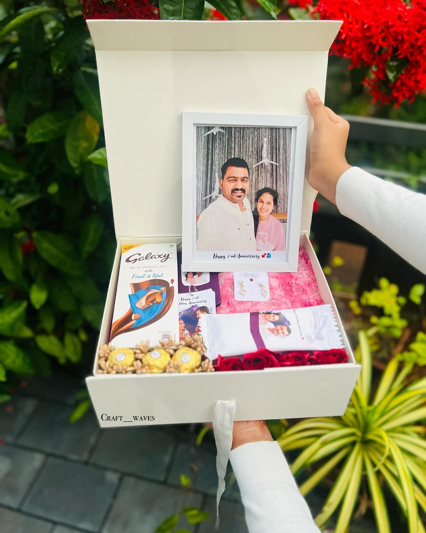 An elegant box featuring a personalised '2nd Anniversary' photo frame, earrings, saree fabric, roses, Galaxy chocolate, and Ferrero Rocher.
