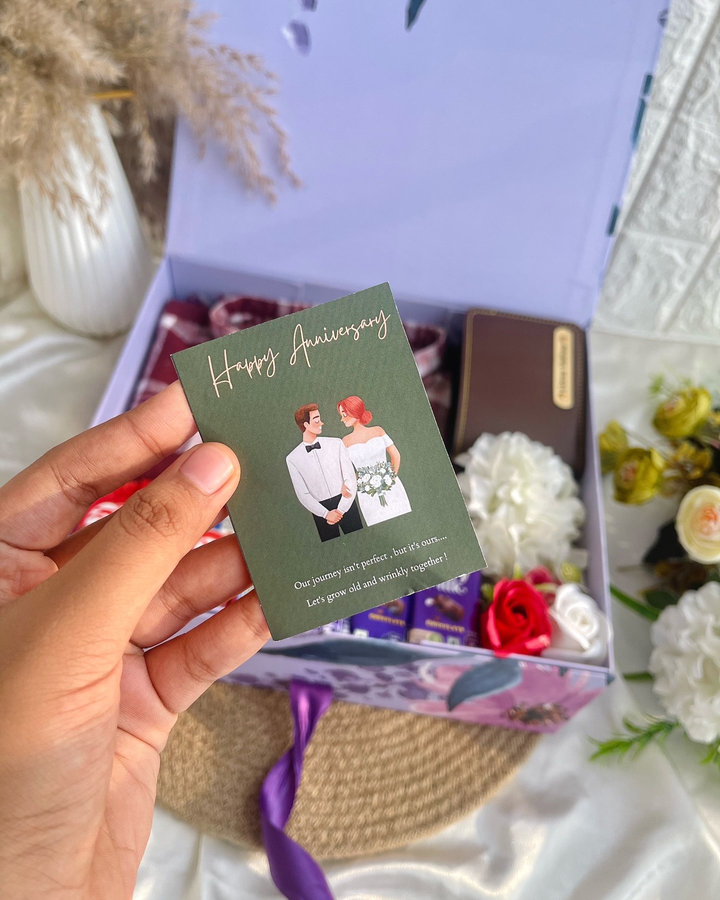 A woven basket gift box holding a leather wallet and assorted chocolates, wrapped with fresh roses and a heartfelt anniversary card.