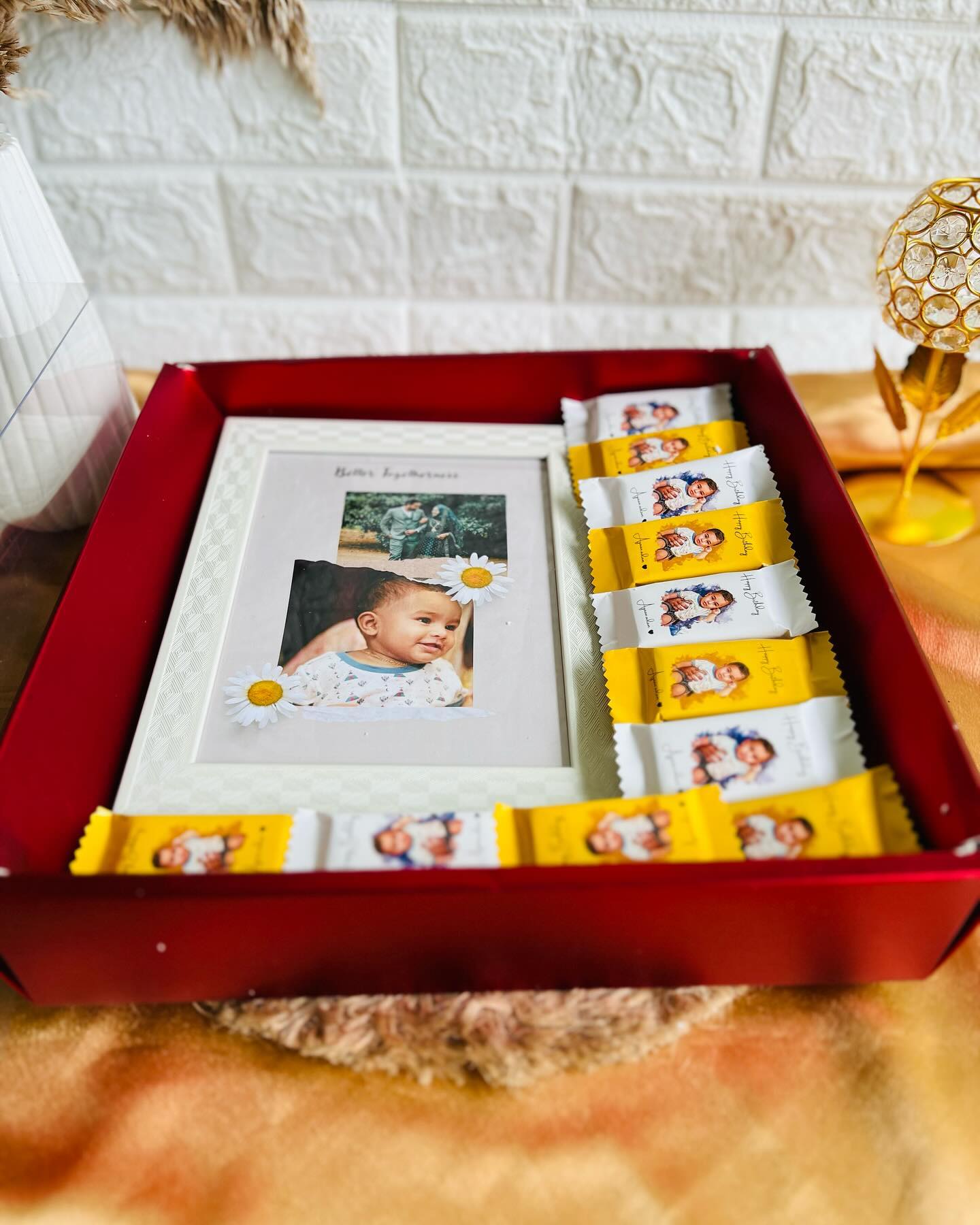 A red gift box with a family photo frame and rows of custom-wrapped 'Happy Birthday' chocolates featuring the baby's photo.