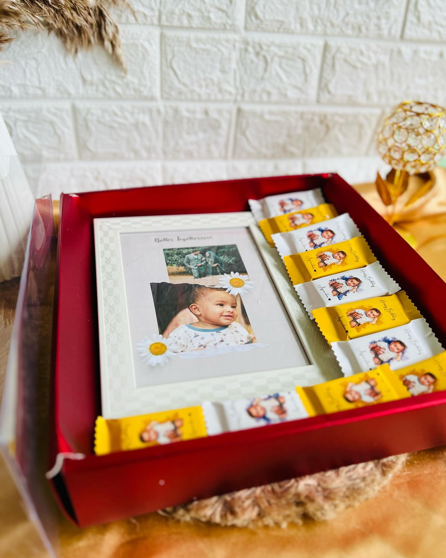 A framed baby photo styled with daisies alongside a row of personalized chocolate bars in cheerful yellow and white wrappers.