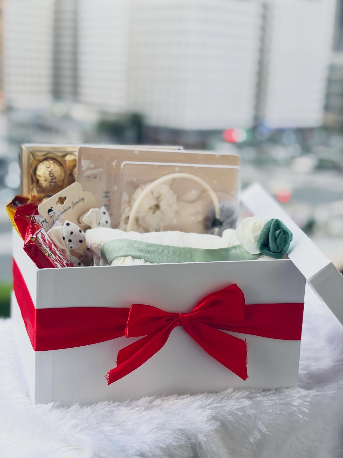 A white gift box tied with a red satin bow, filled with soft baby accessories, chocolates and floral touches to welcome a new little one.