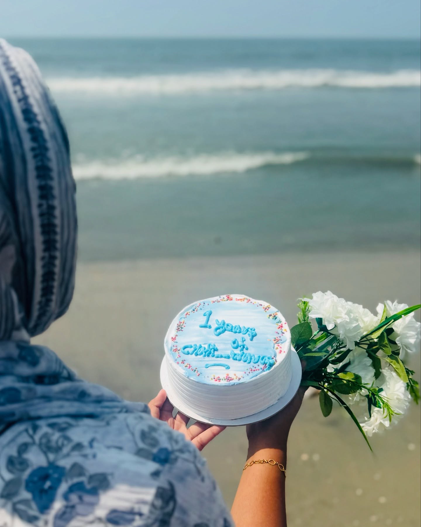 A white buttercream cake piped with a custom milestone message and sprinkles, paired with white blooms by the shore.