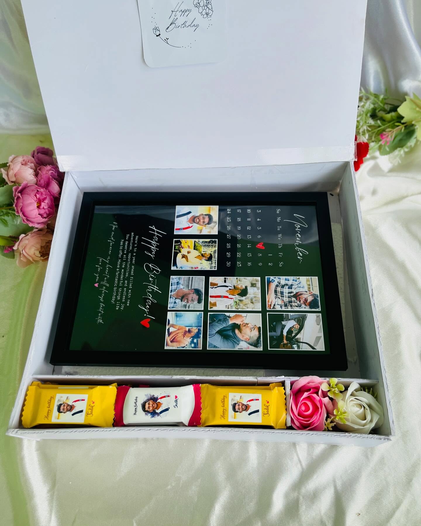 A gift box featuring a personalized calendar-style photo frame, chocolate bars and a handwritten birthday card.