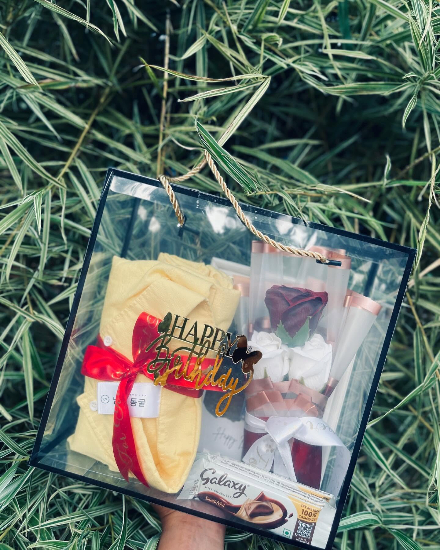 A clear gift bag with a folded yellow outfit, dried rose bouquet, Galaxy chocolate, and a gold Happy Birthday topper.