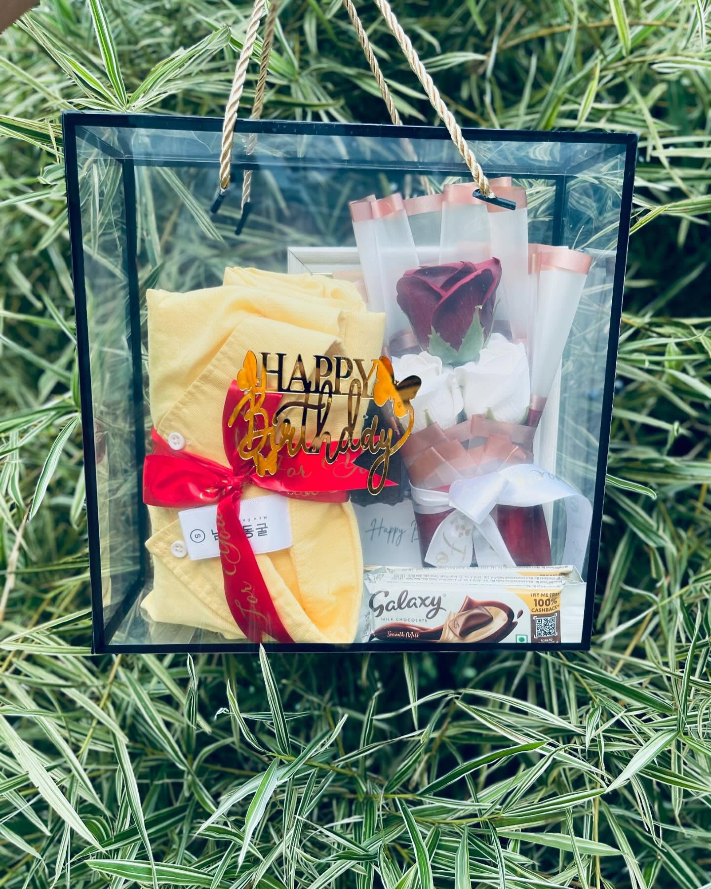 A hanging glass gift box combining a folded shirt, a gold 'Happy Birthday' topper, rose bouquets, and Galaxy chocolate.