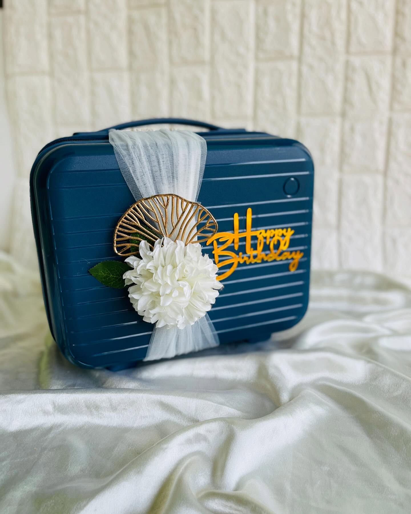 A navy hard-shell mini suitcase gift box dressed with a tulle bow, white bloom, and gold 'Happy Birthday' topper.