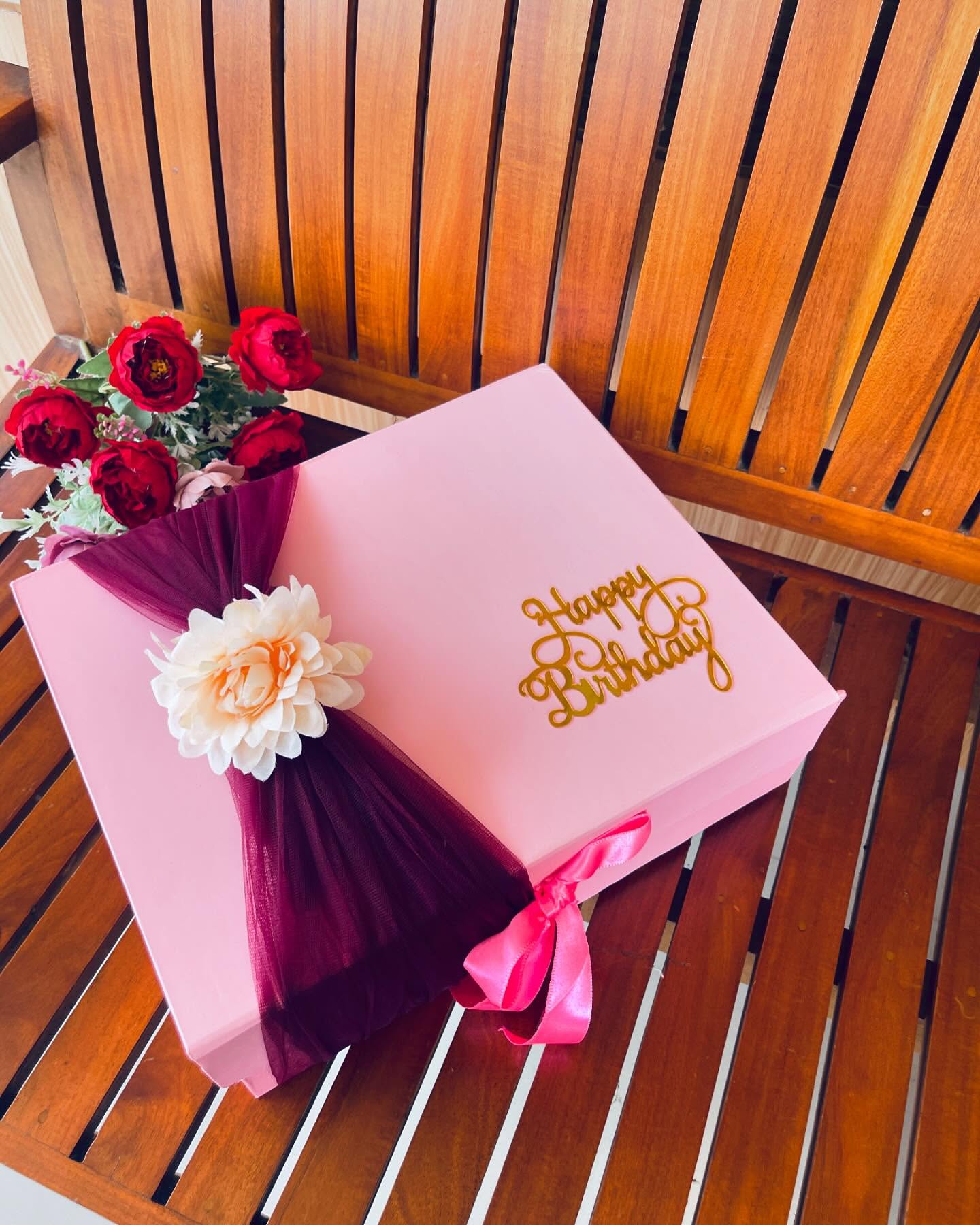 A blush pink gift box wrapped with a deep burgundy tulle sash, a cream bloom accent, and a gold 'Happy Birthday' topper.