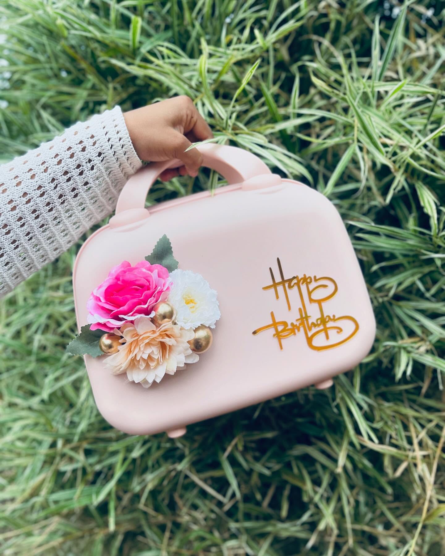 A blush pink vanity case decorated with silk florals and a gold 'Happy Birthday' script, doubling as a keepsake bag.