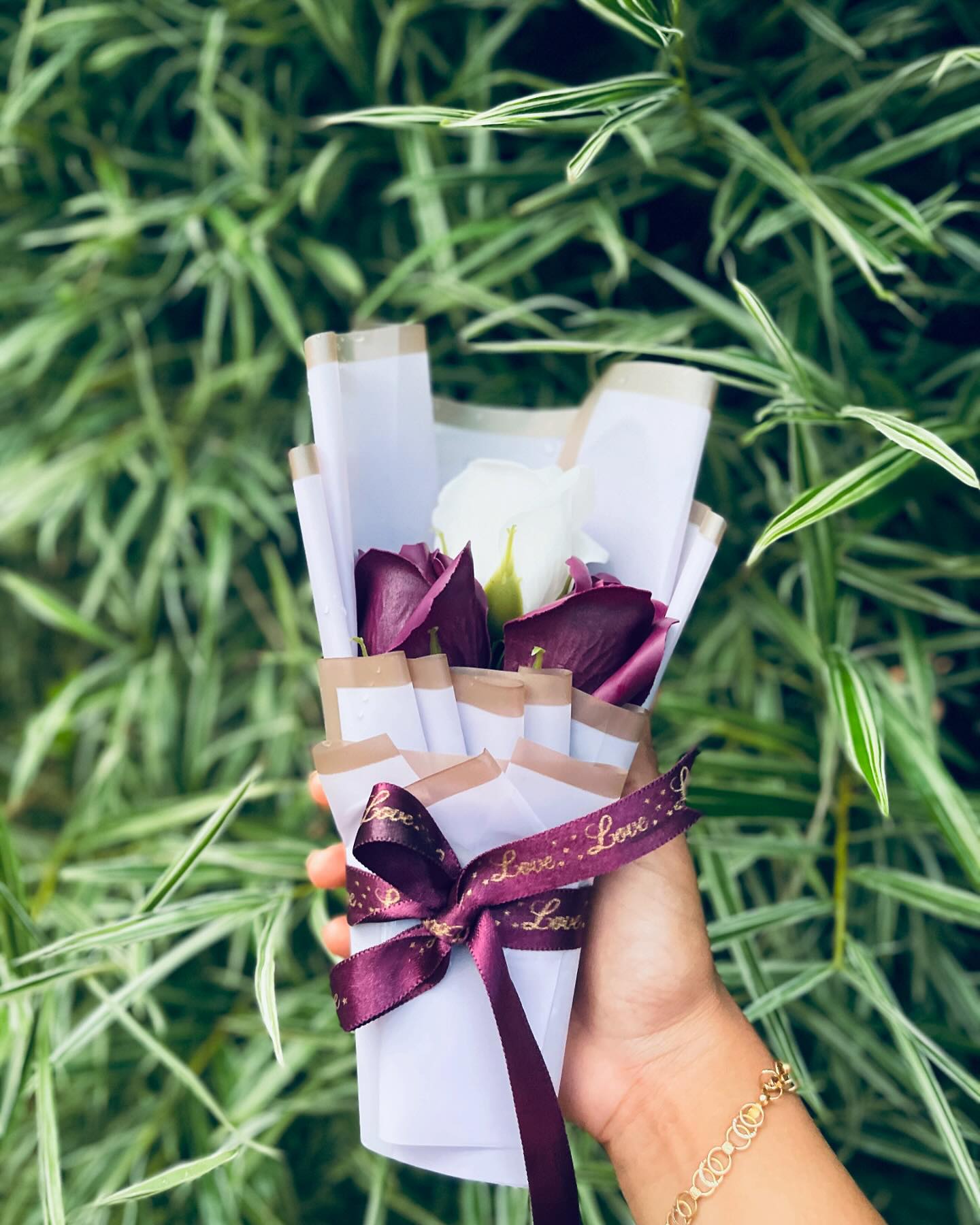 A romantic hand-tied bouquet of deep burgundy and ivory roses wrapped in white paper with a 'Love' satin ribbon.
