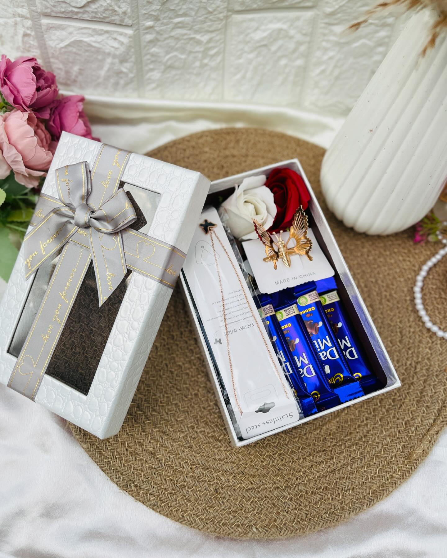 A white ribboned gift box with a stainless steel necklace, a gold butterfly hair clip, silk roses and Dairy Milk chocolates.