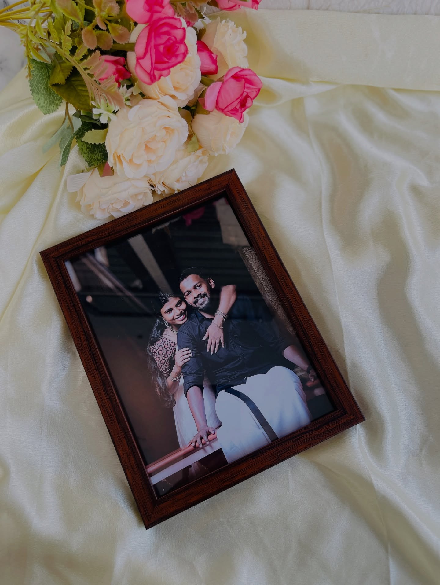 A warm wood-toned photo frame showcasing a candid couple portrait, styled with pink and cream roses.