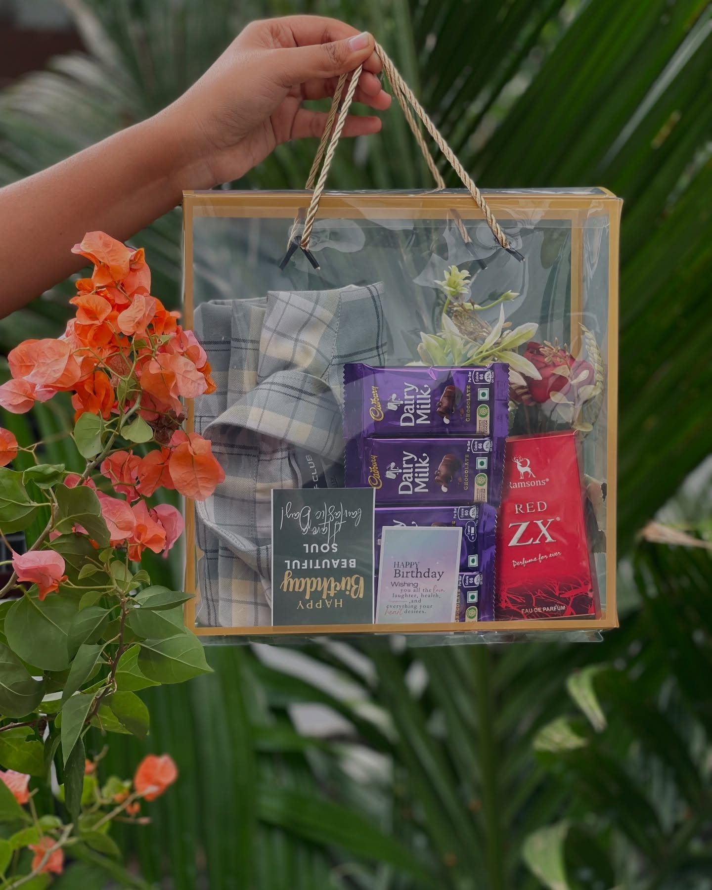 A clear gift bag with a grey checked shirt, Cadbury Dairy Milk bars, a Ramsons perfume, and a birthday card.