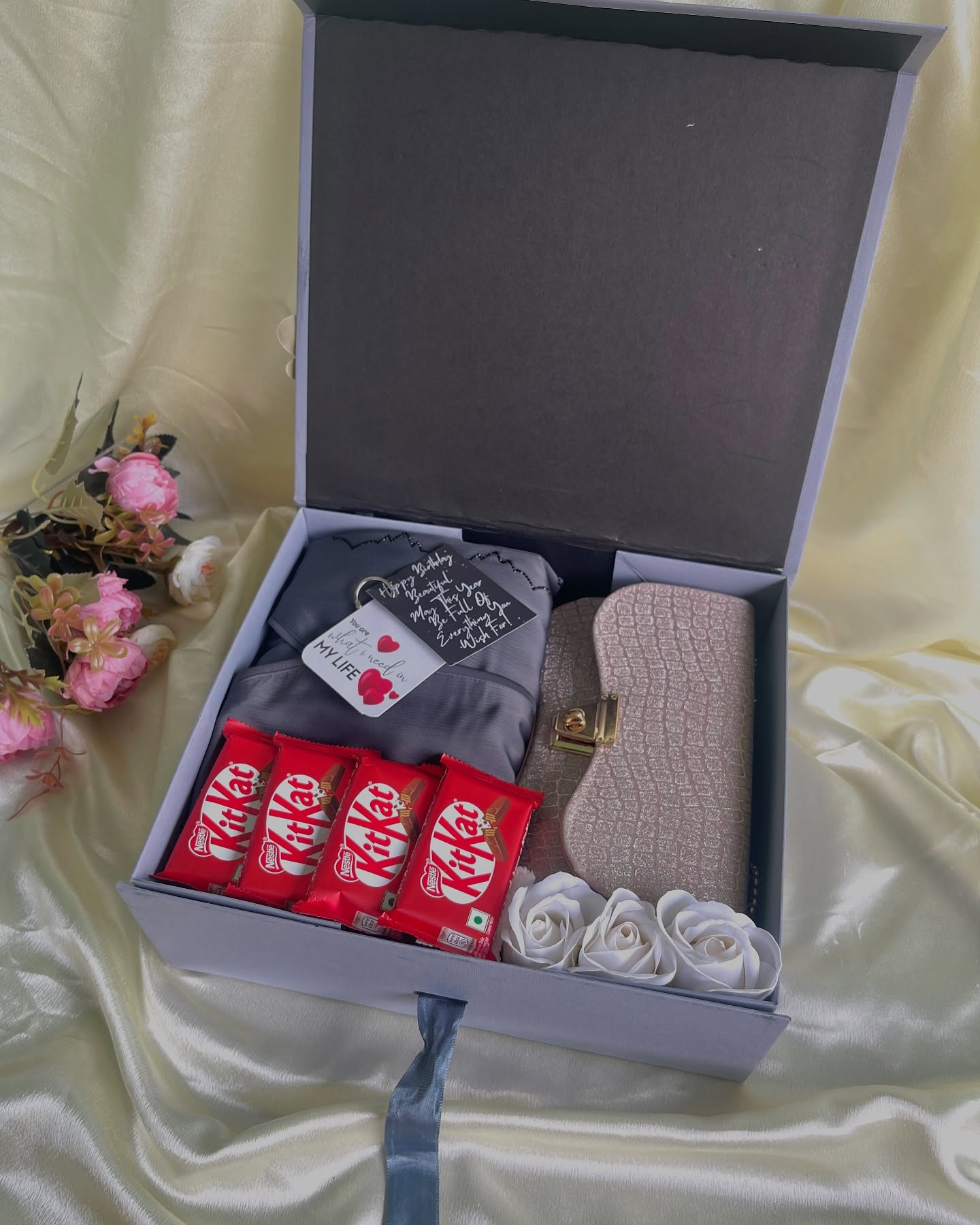 A grey birthday gift box featuring a glittery clutch purse, a cosy pouch, KitKat bars, and soft white roses.