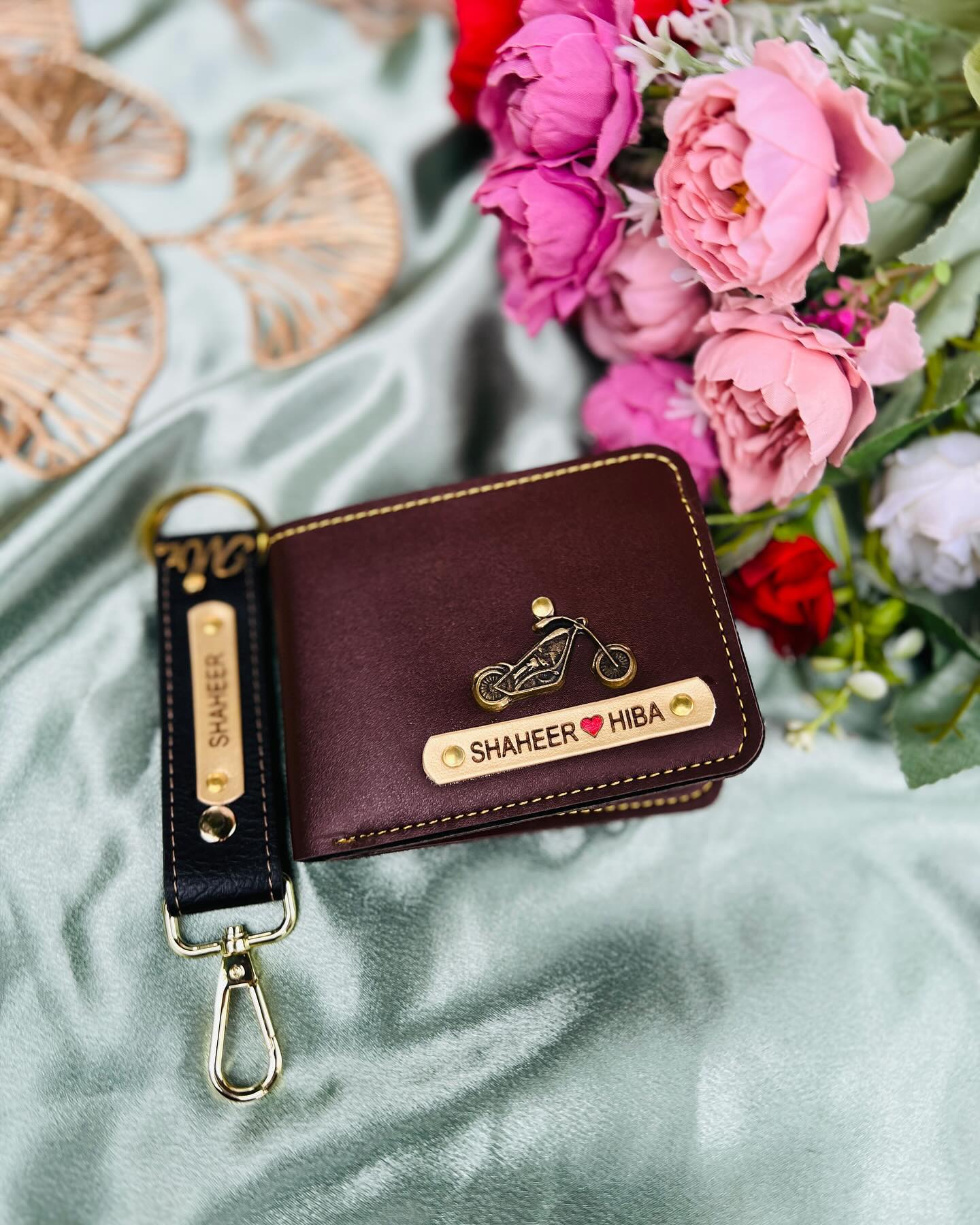 A burgundy leather wallet and matching keychain engraved with both names and a heart, wrapped with fresh blooms.