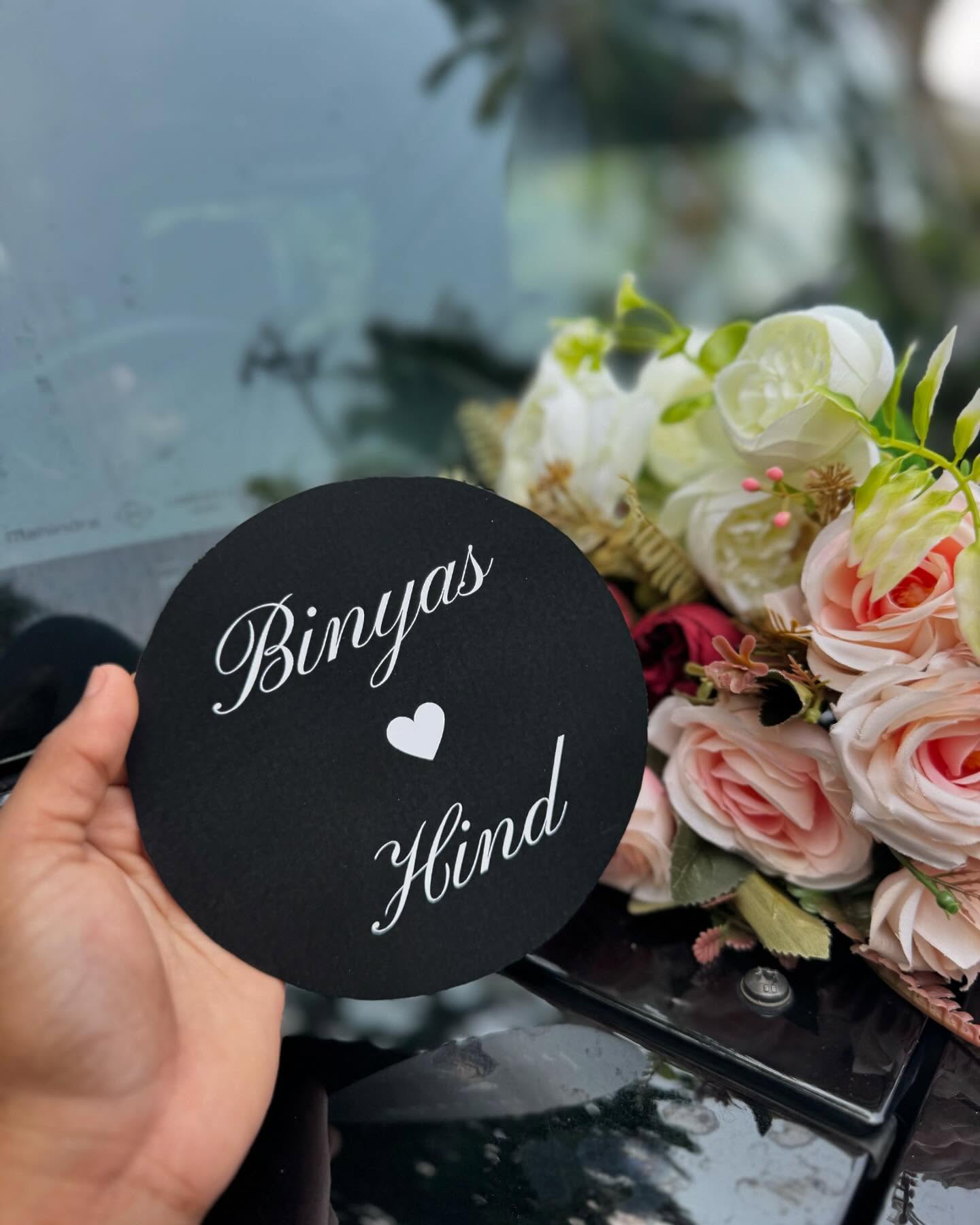 A round black plaque personalized with a couple's names and a heart, set among a floral bouquet backdrop.