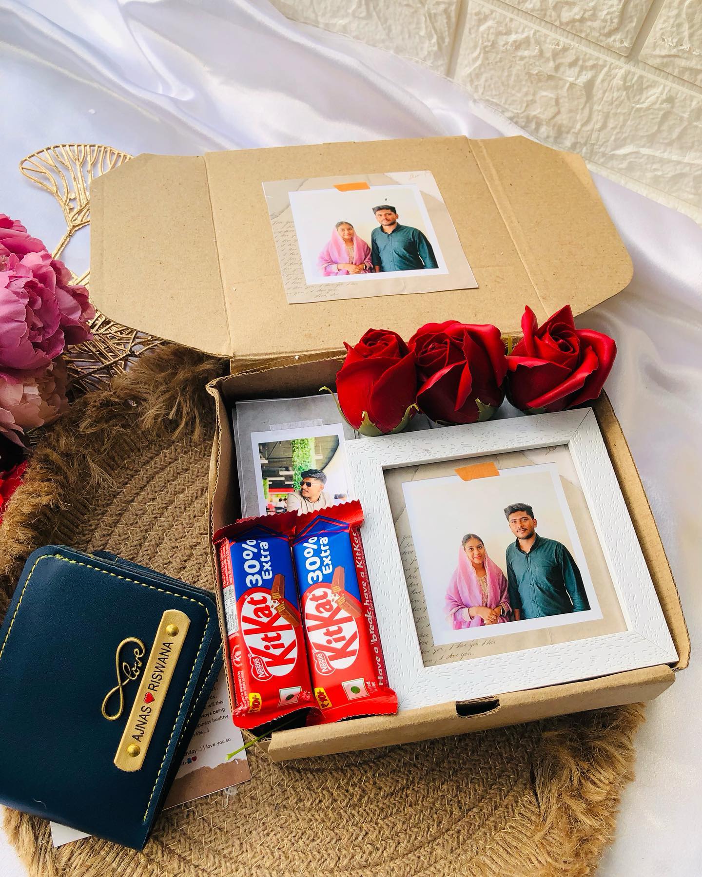 A personalized anniversary box with framed couple photos, red roses, KitKat bars, and a custom-engraved wallet.