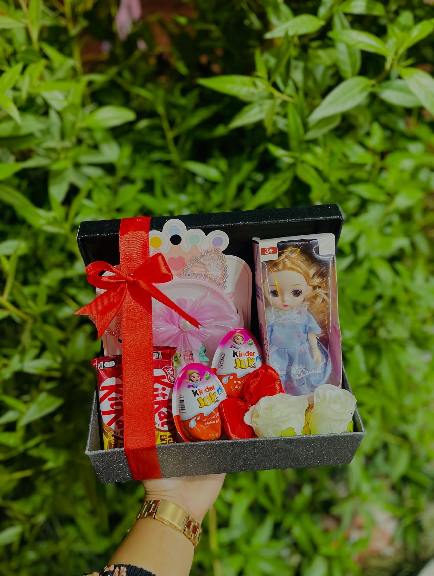 A playful kids' gift box with a dress-up doll, Kinder Joy eggs, KitKat, and soft roses, finished with a red satin bow.