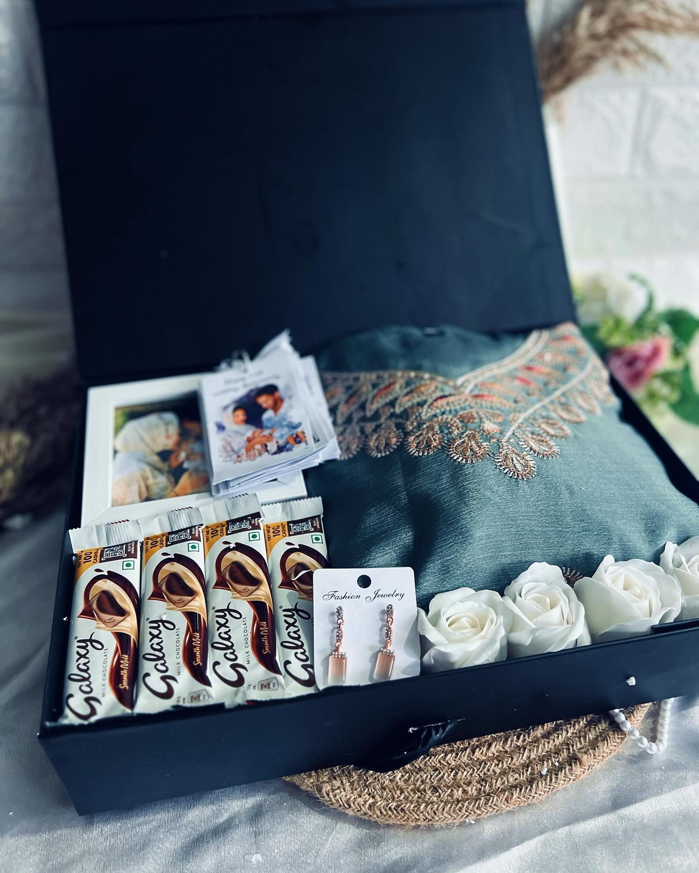An open black gift box pairing an embroidered fabric piece with chocolates, earrings, and a framed keepsake photo.
