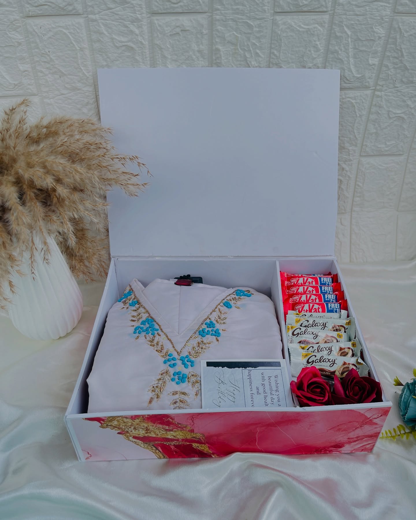 An open gift box featuring a blue-embroidered white kurta alongside KitKat and Galaxy chocolates, roses and a birthday card.