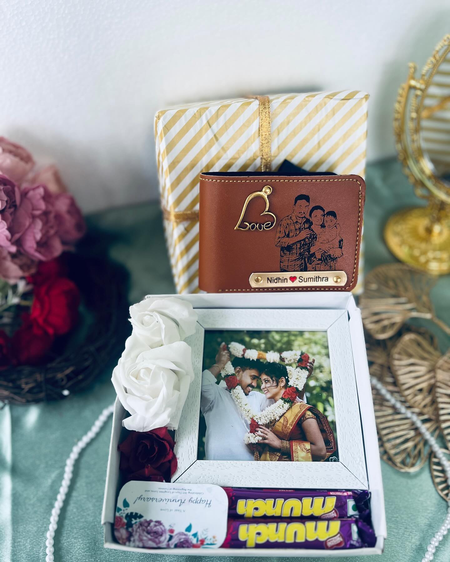 A gold-striped gift box pairing a personalized engraved leather wallet with a framed wedding photo, soft roses, and Munch chocolates.