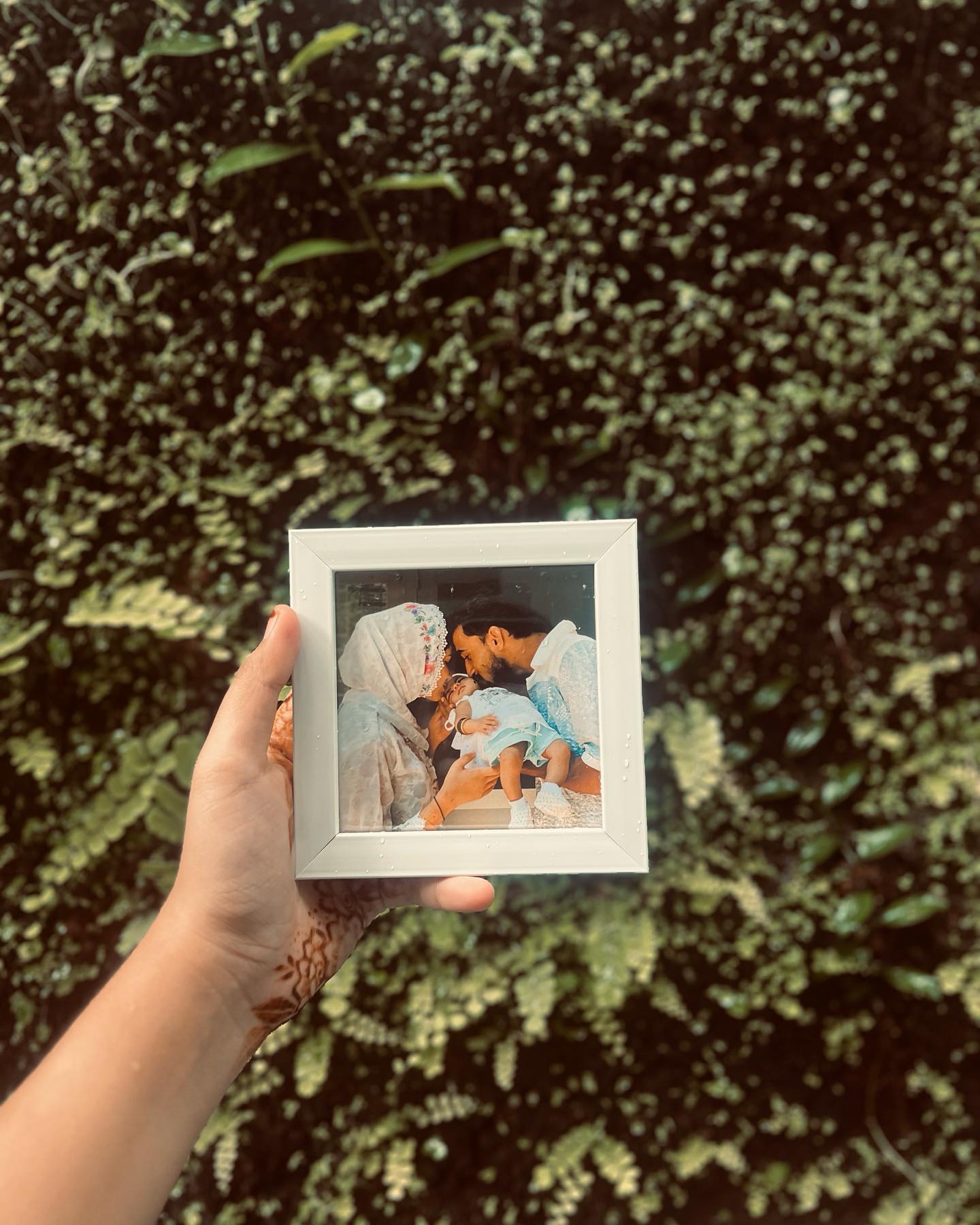 A natural wood photo frame printed with a tender family portrait, held outdoors against greenery.