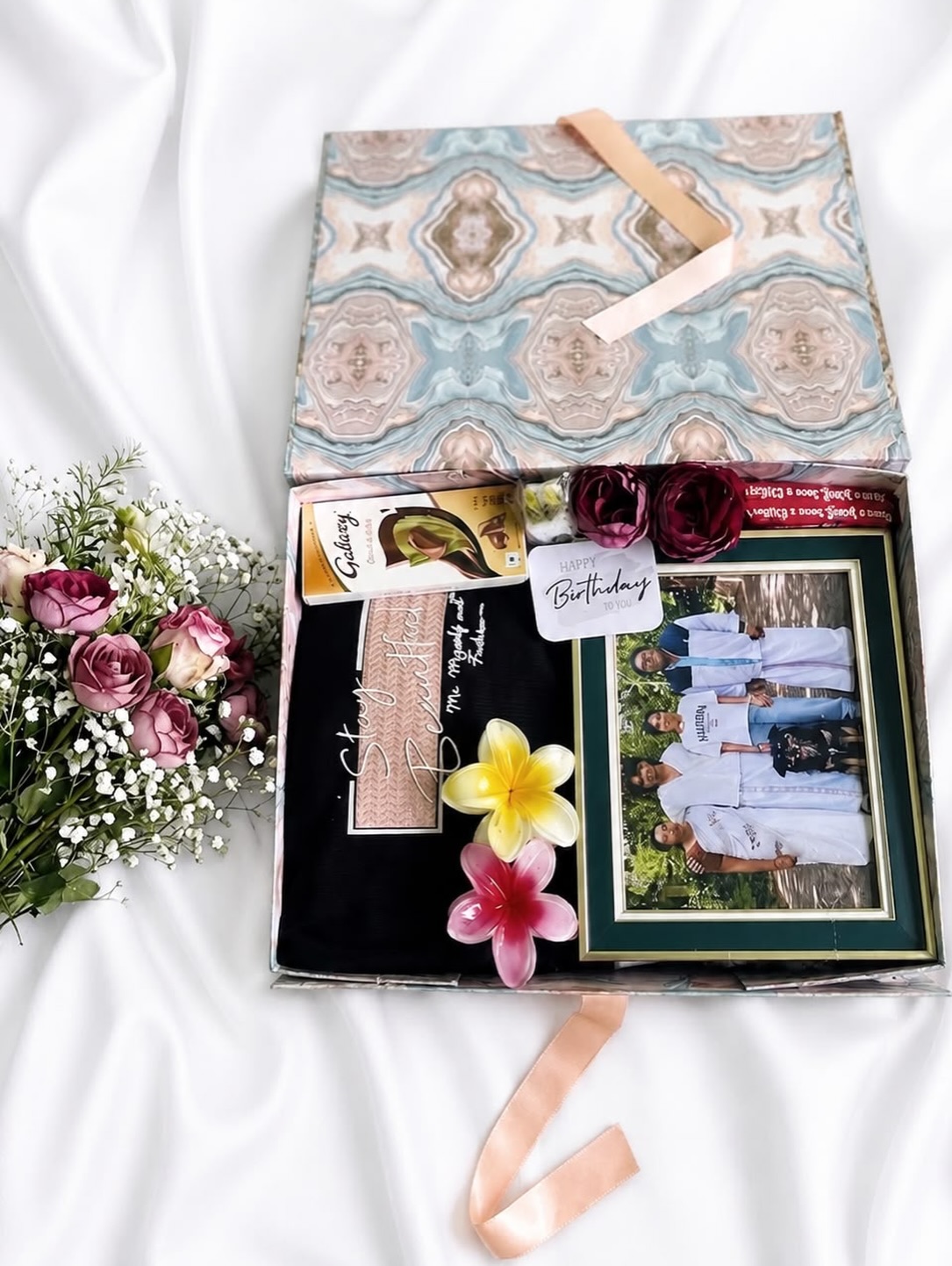 A patterned birthday gift box with a framed family photo, Galaxy chocolate, a scarf, and tropical flower accents.