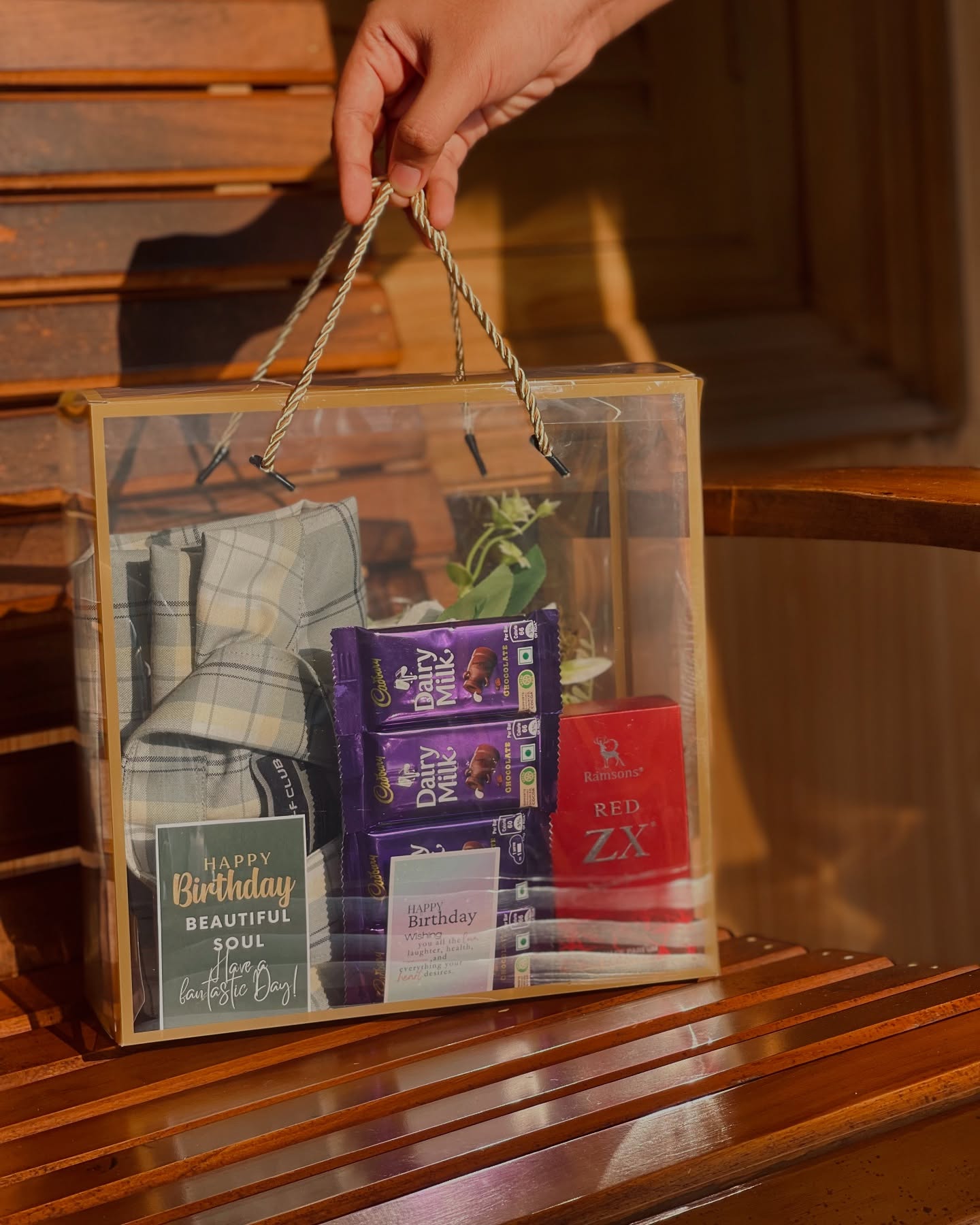 A clear gift bag with a checkered flannel shirt, Cadbury chocolates, a fragrance box, and a 'Happy Birthday' card.