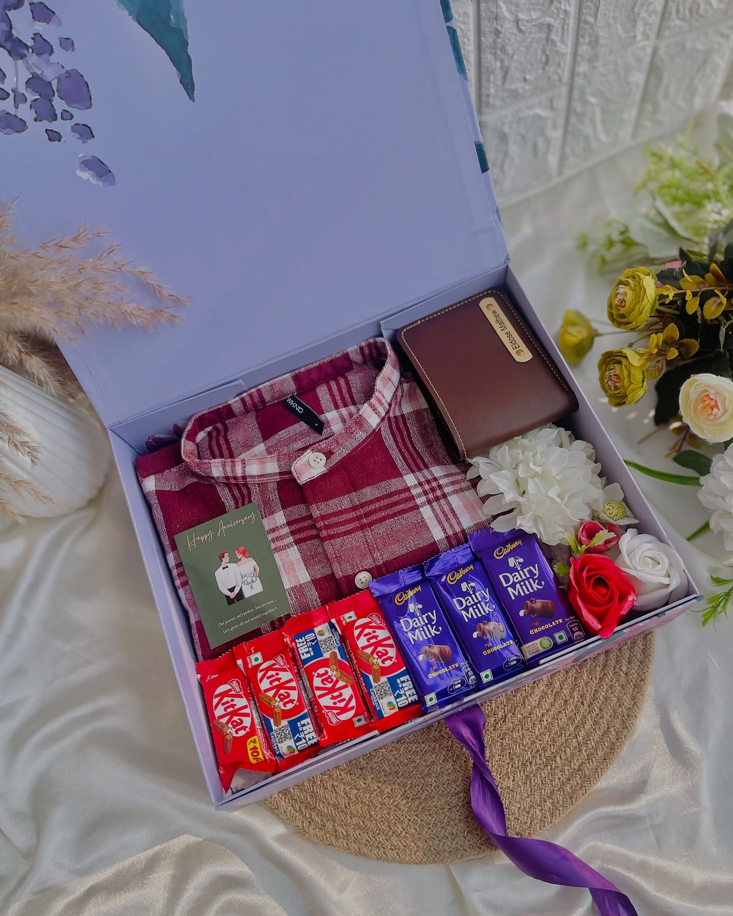 An anniversary gift box for him with a checked flannel shirt, a name-engraved wallet, chocolates and roses.
