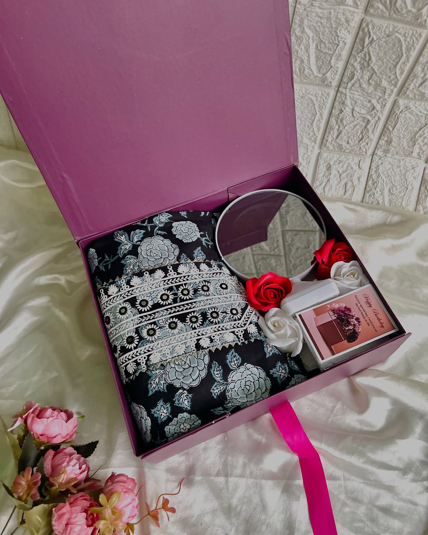 A plum gift box featuring an embroidered floral saree, a round vanity mirror, red roses, and a birthday card.