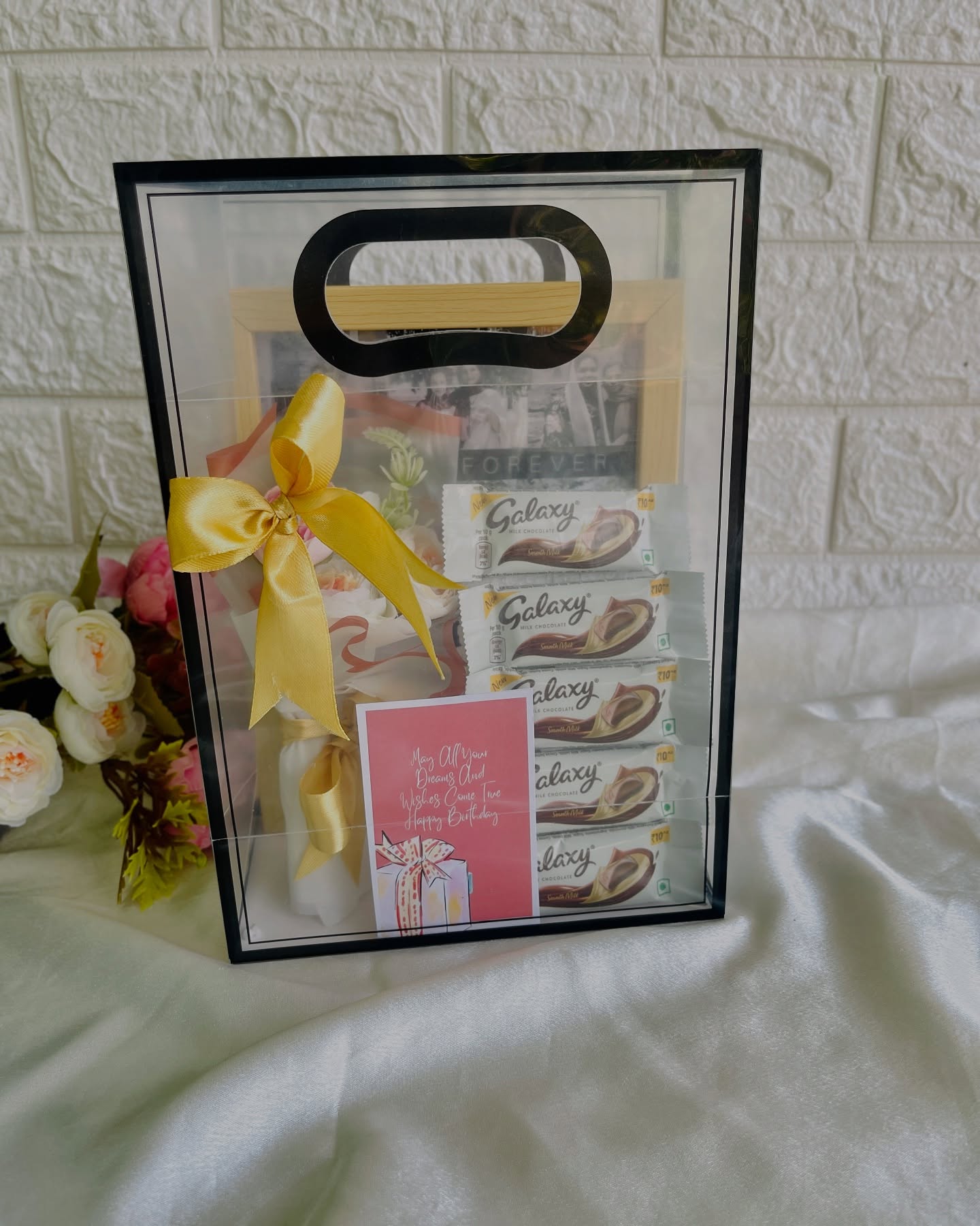 A clear gift bag featuring a personalized 'Forever' photo frame, fresh flowers, Galaxy chocolates, and a birthday card.