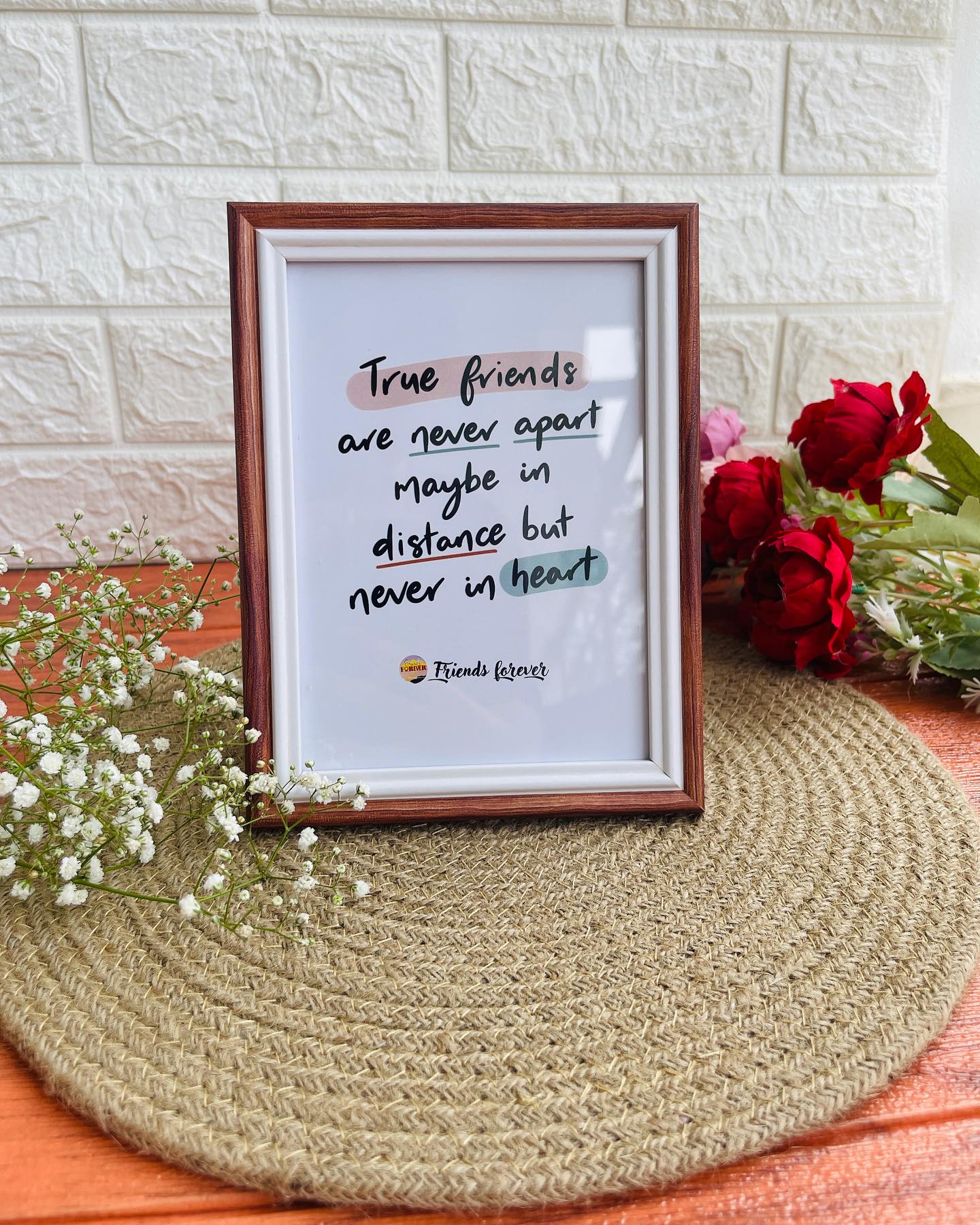 A hand-lettered "true friends are never apart" quote print in a wooden frame, styled with roses and baby's breath.