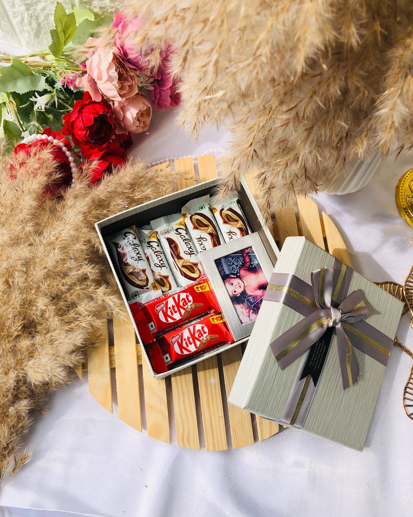 An open keepsake box layered with Galaxy and KitKat chocolate bars beside a mini printed photo, tucked into a ribboned gift box.