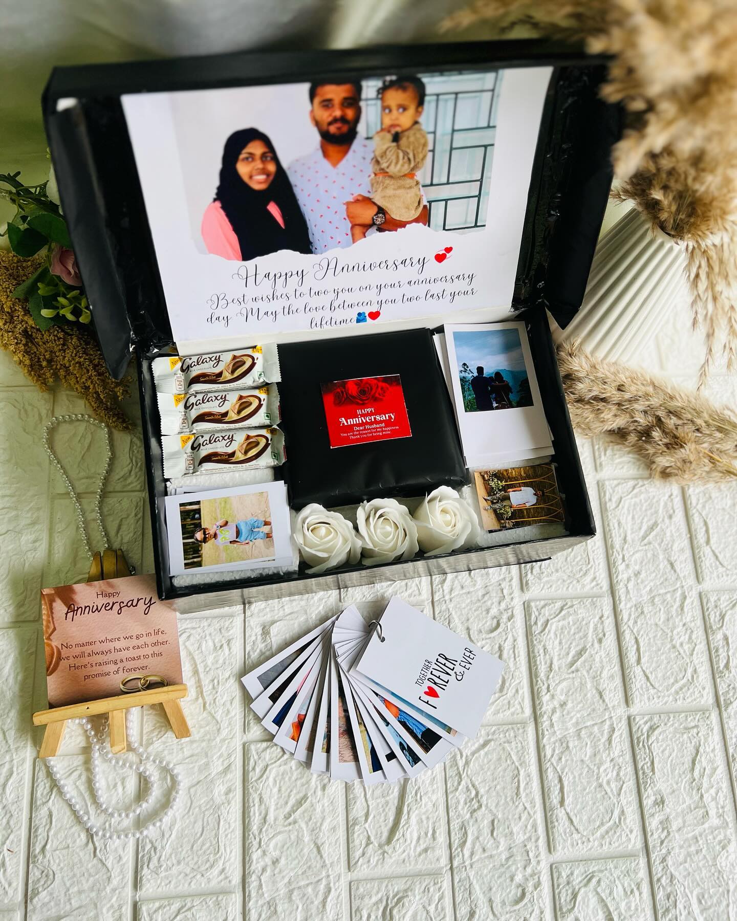 An elaborate anniversary keepsake box with Galaxy chocolates, foam roses, a printed couple photo lid, and a fan of mini memory cards.