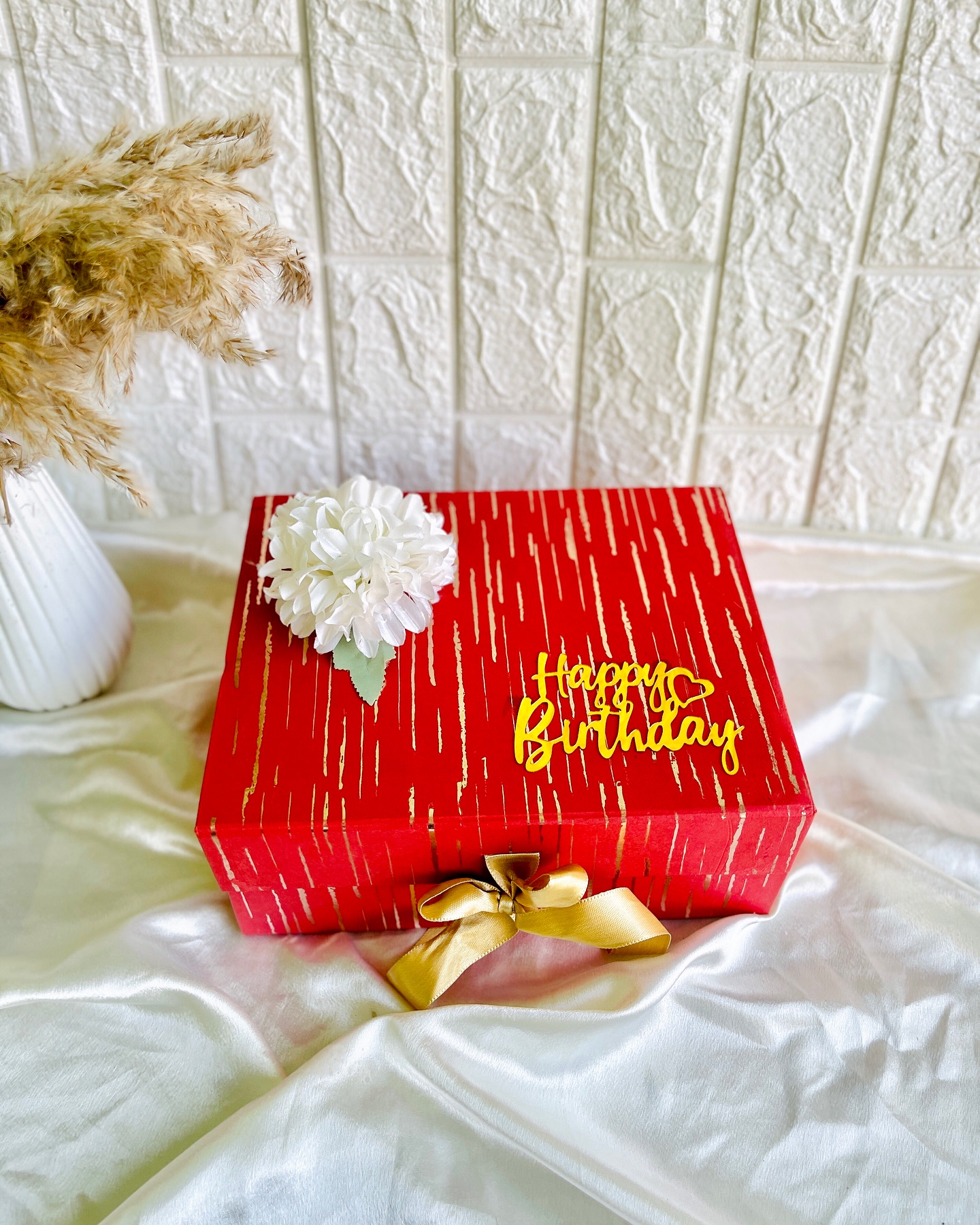 A red and gold-striped birthday gift box topped with a white bloom and a gold Happy Birthday charm.