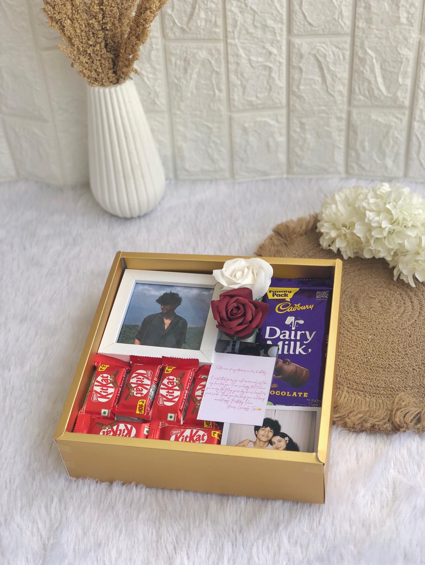 A gold gift box with a personalized photo frame note, family photos, silk roses, KitKats and a Dairy Milk bar.