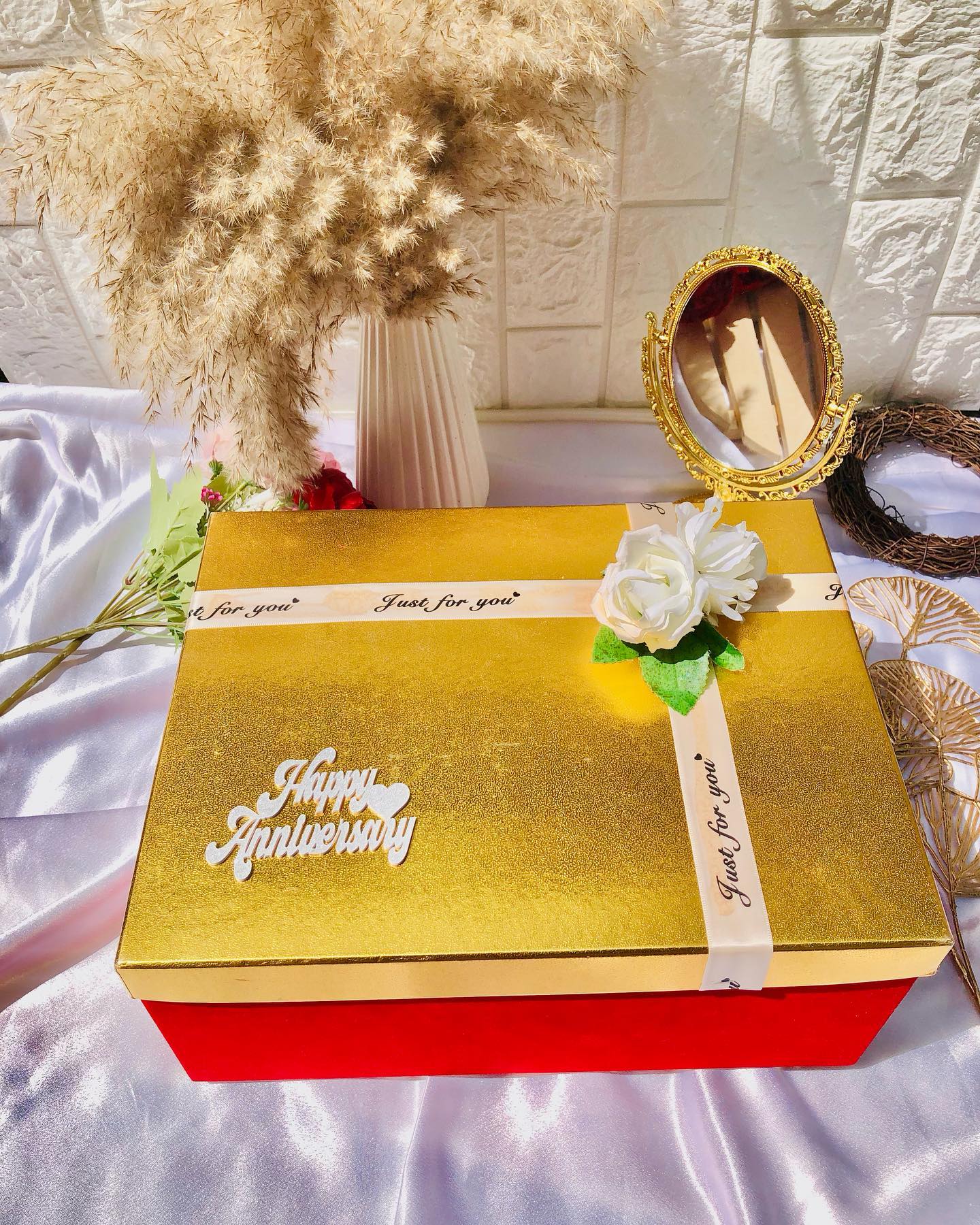 A shimmering gold gift box with a "Happy Anniversary" topper, ribbon, and white rose accent.