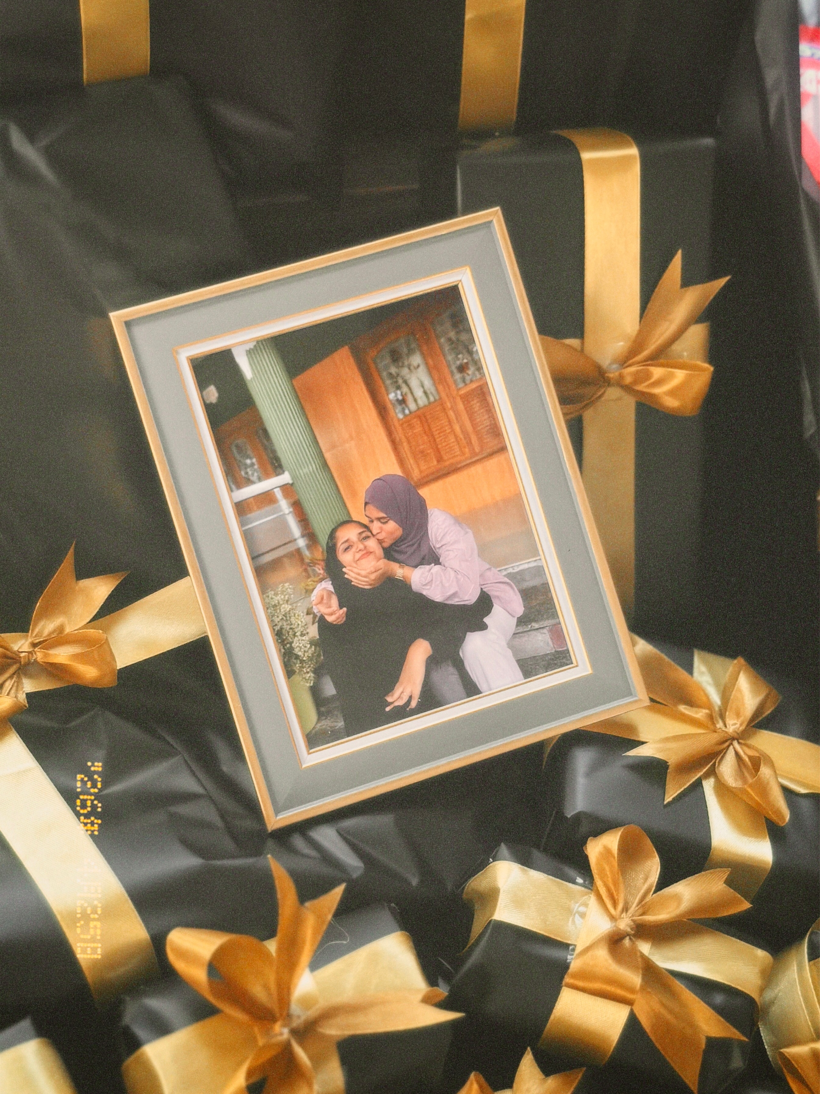A gold-edged photo frame printed with a romantic couple portrait, styled amid black gift boxes wrapped in gold ribbon.