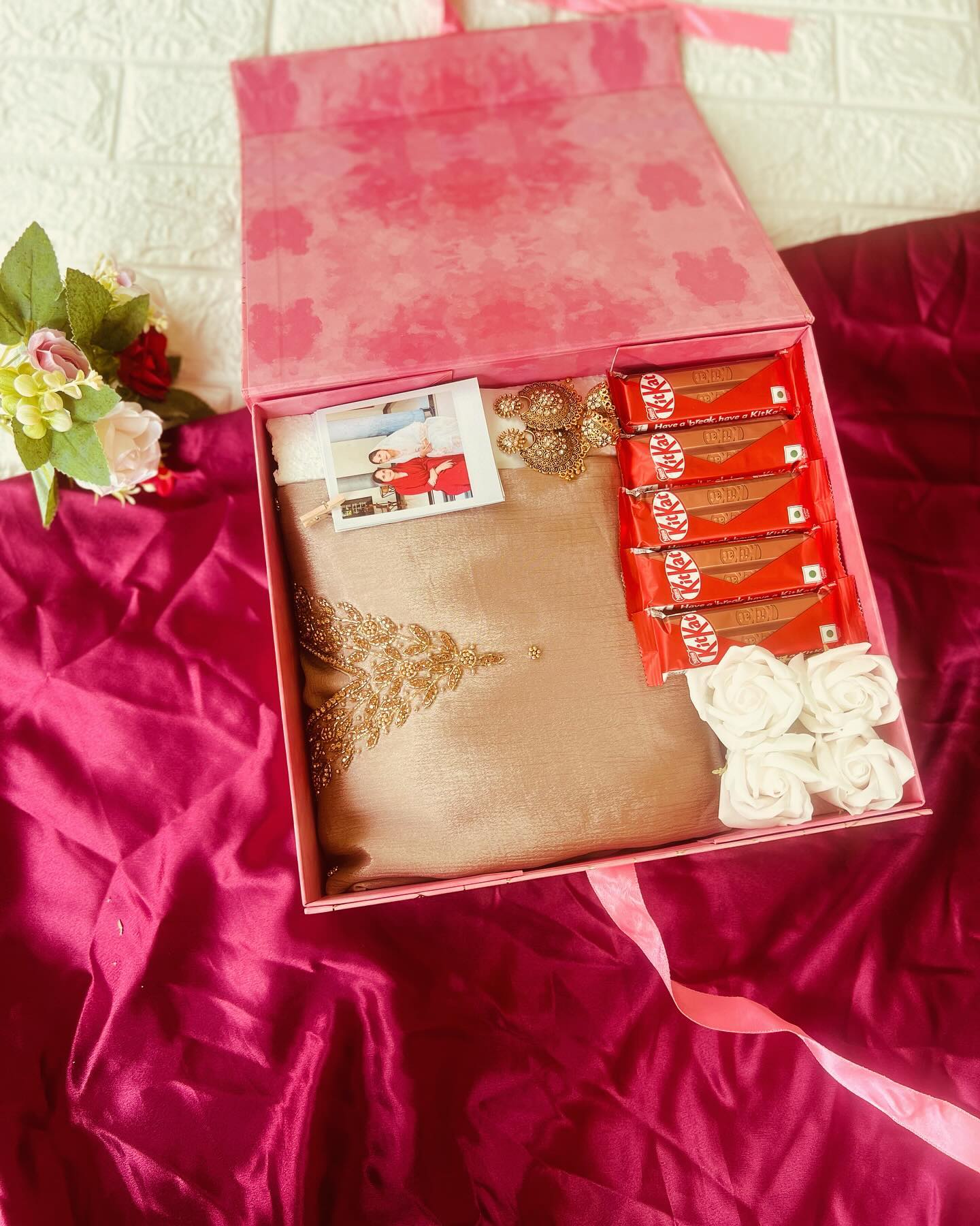 A pink gift box layering a beige embellished saree, gold-toned jewelry, KitKat bars, and a personalized photo.