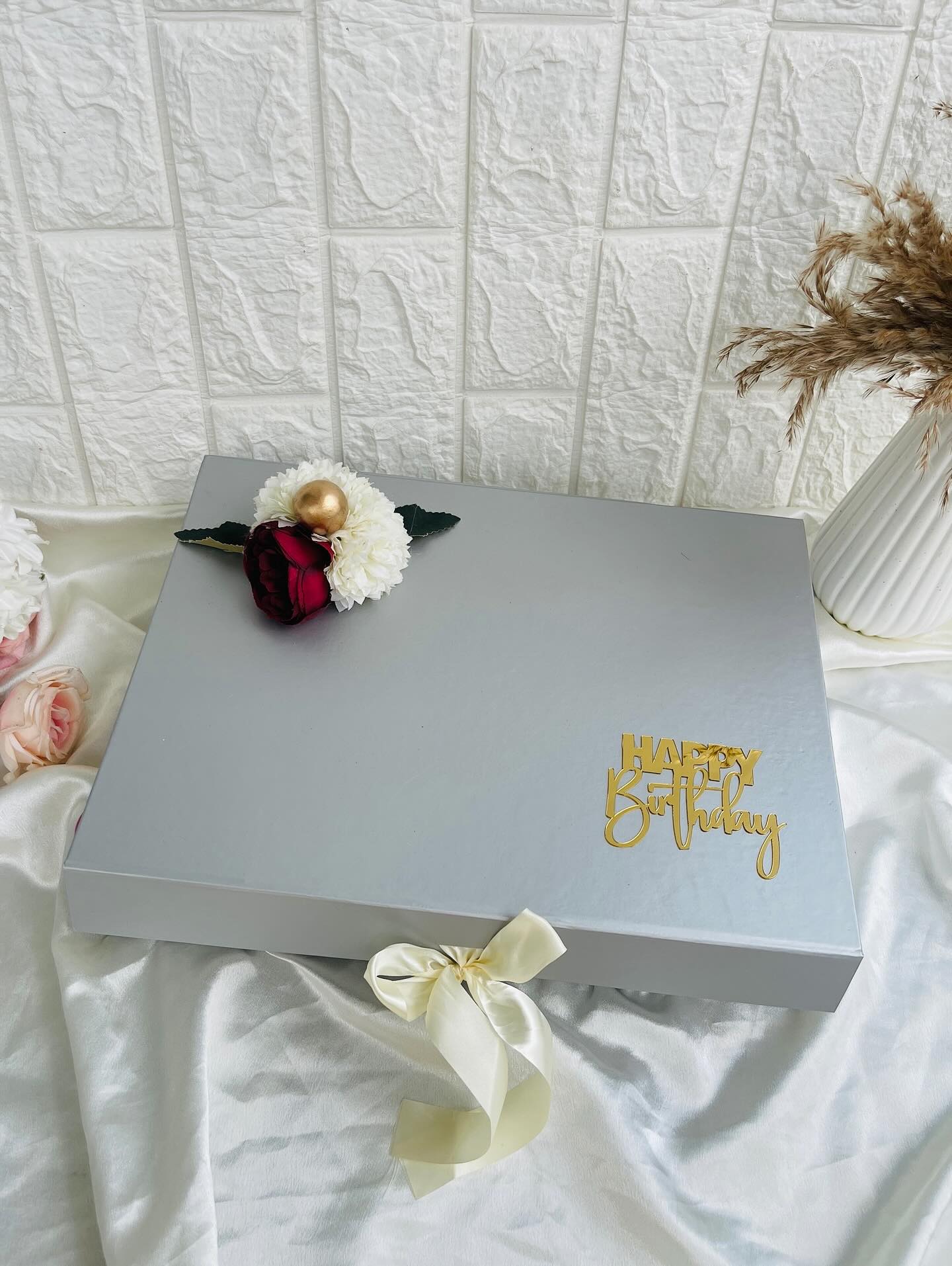 A sleek slate-grey gift box finished with a fresh floral topper, ivory satin ribbon, and a gold Happy Birthday sign.