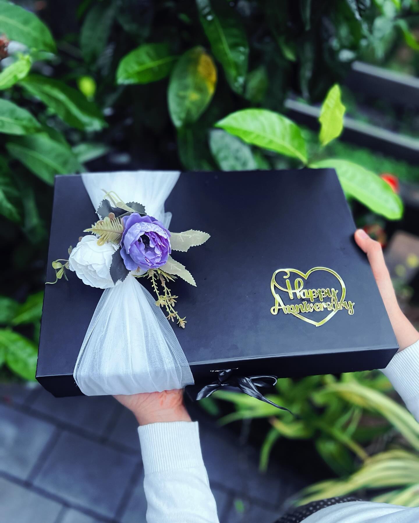 An elegant black gift box adorned with a gold heart "Happy Anniversary" seal and tulle florals.