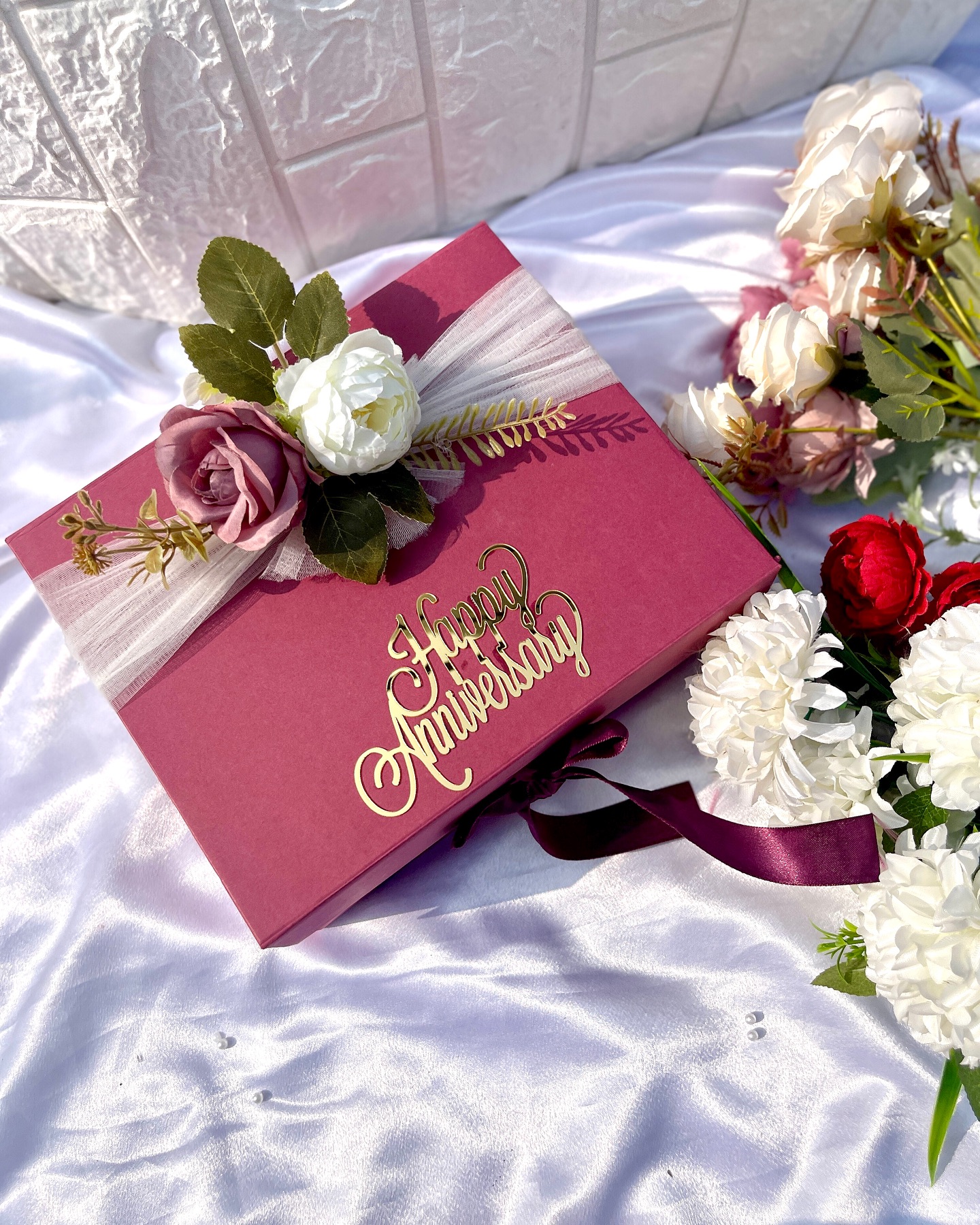 A rose-pink gift box with a gold 'Happy Anniversary' script topper and a fabric rose and tulle bow.