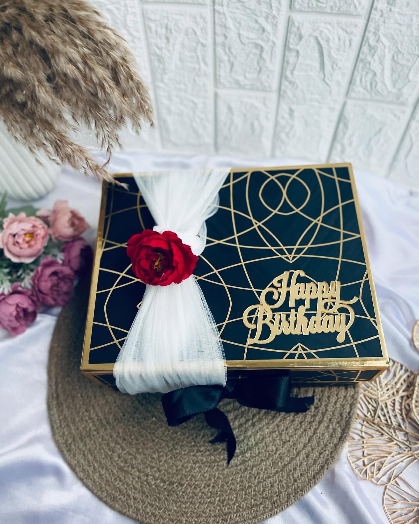 A sleek black and gold geometric gift box wrapped with white tulle, a red rose and a black satin bow.