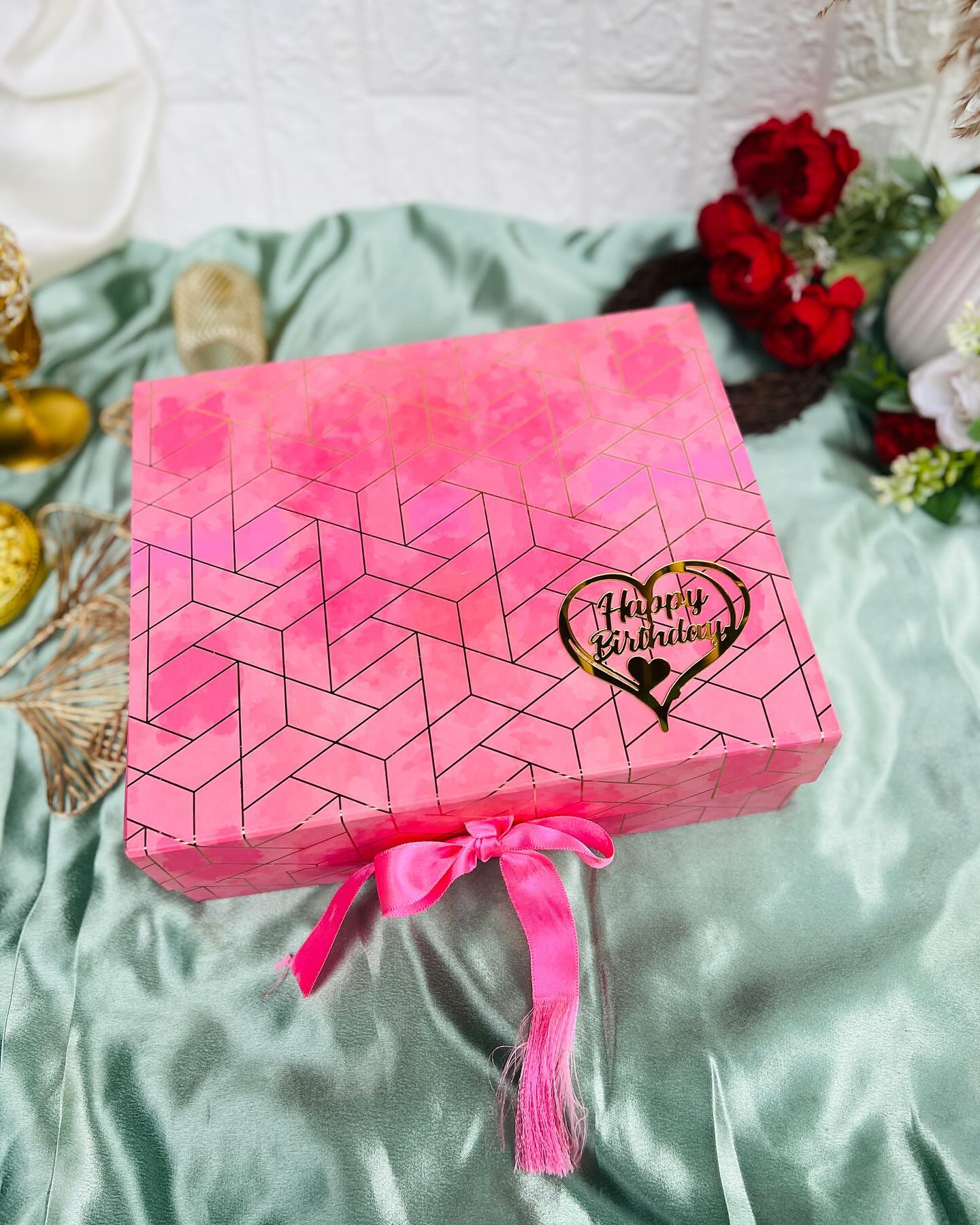 A pink geometric-patterned gift box with a mirrored gold "Happy Birthday" heart charm and a pink satin bow.