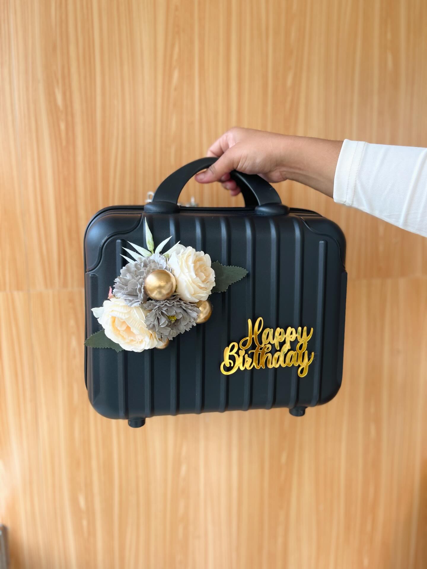 A chic matte-black mini suitcase gift box topped with a cream rose and gold berry arrangement and a gold 'Happy Birthday' charm.