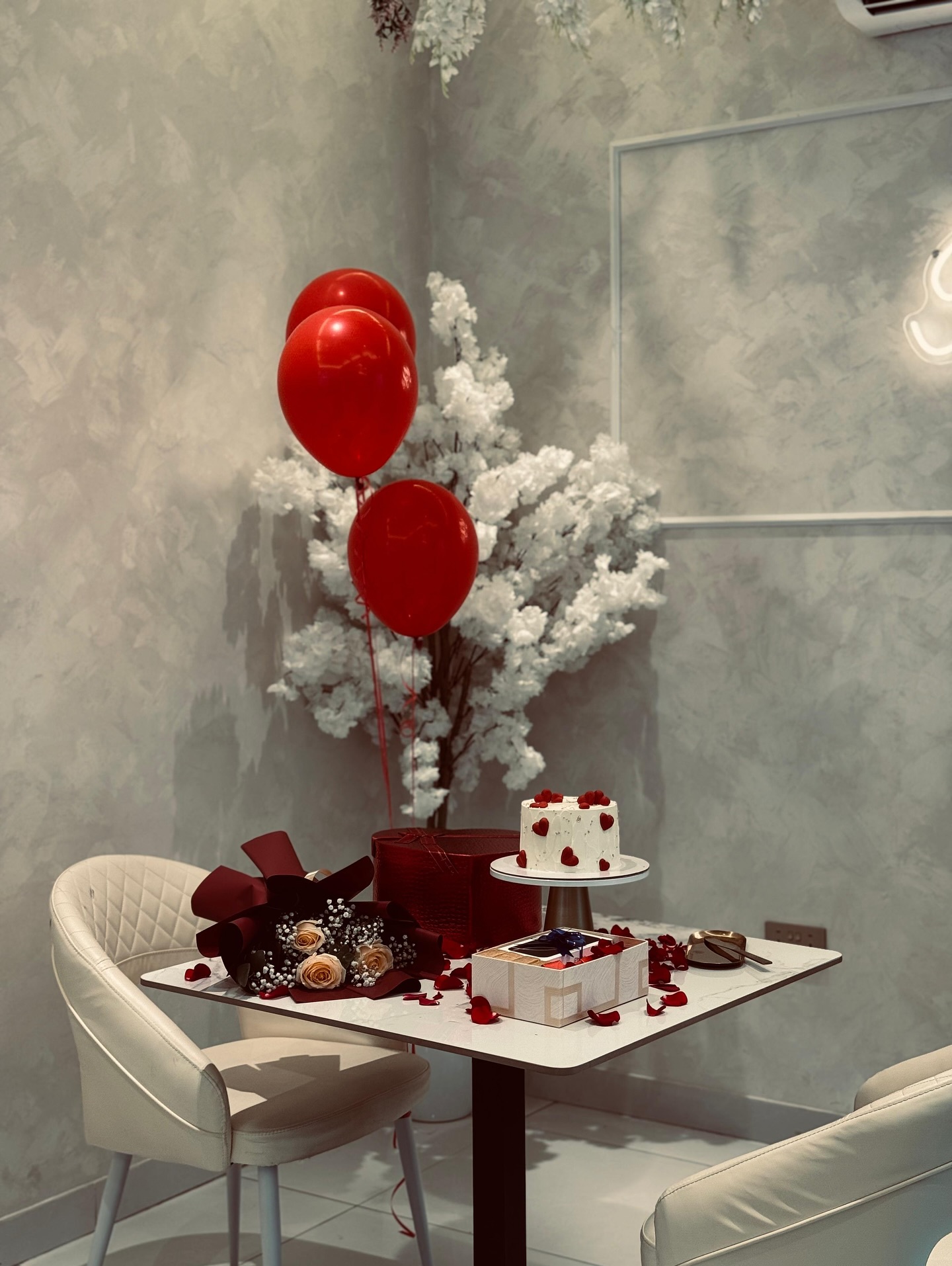 A red-heart-decorated celebration cake paired with a yellow rose bouquet and gift box, set against balloons and florals.