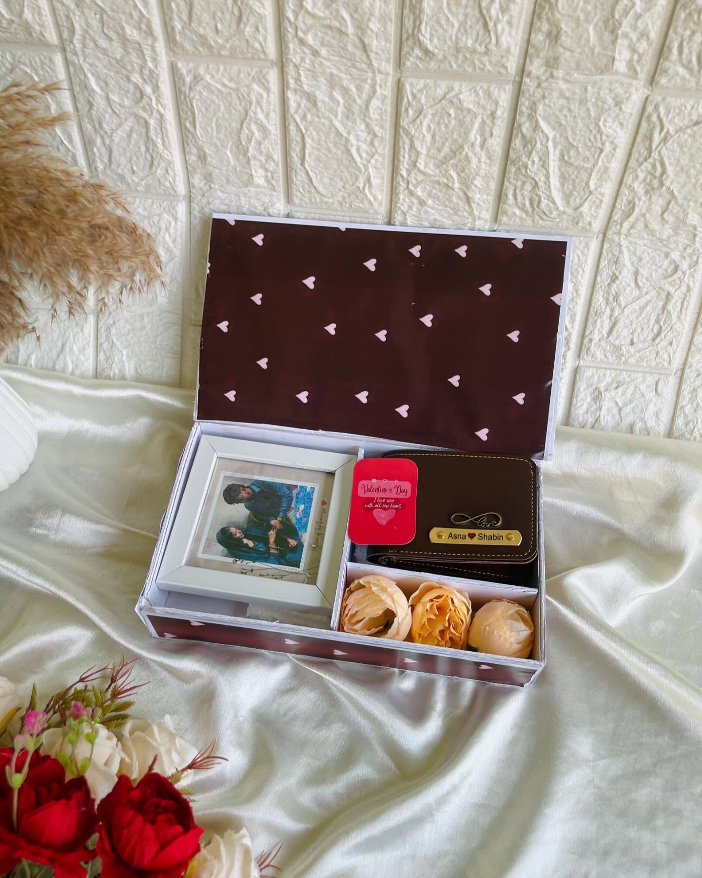 A heart-patterned gift box with a couple photo frame, a personalized dual-name wallet, and soft peach roses.