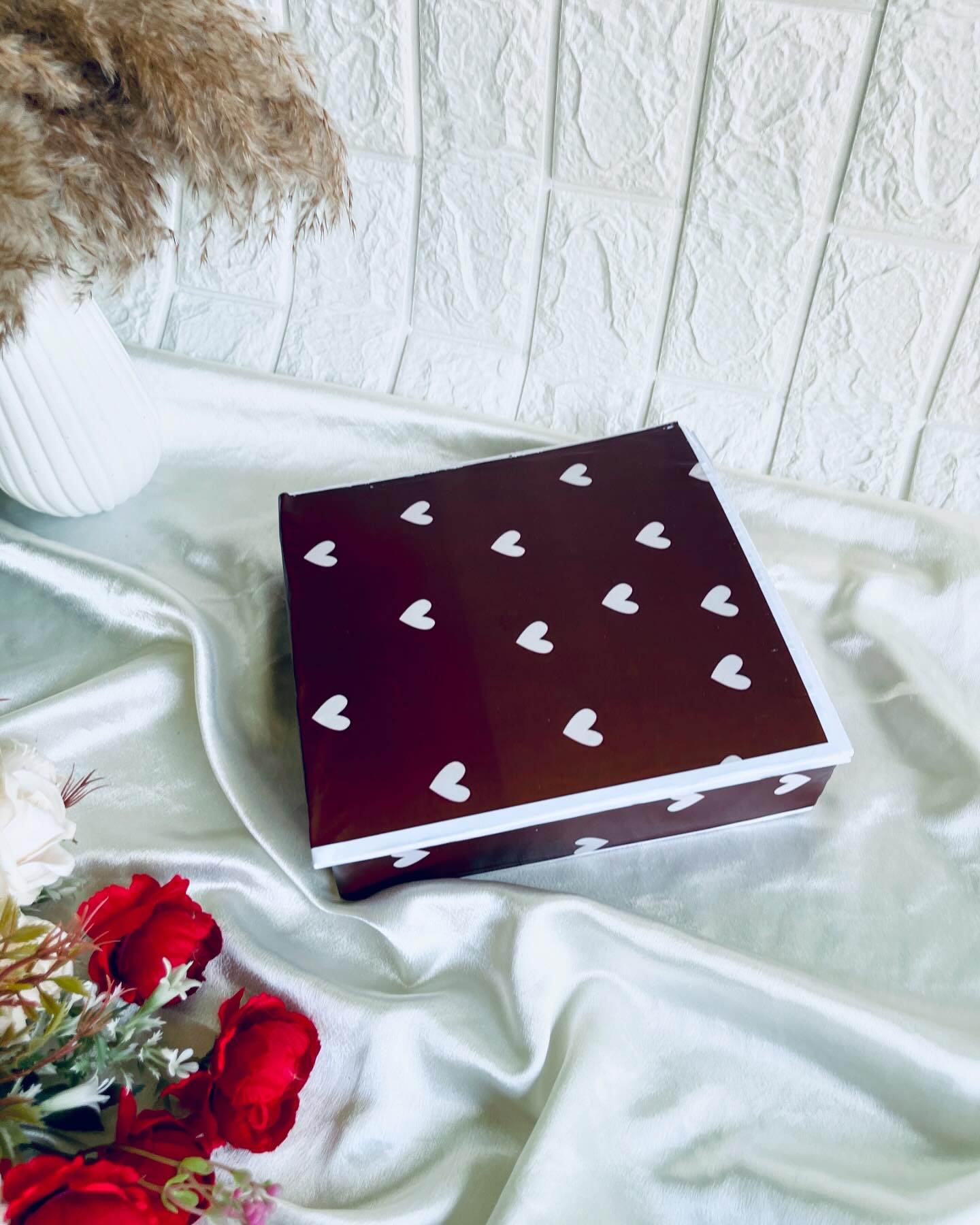 A compact chocolate-brown gift box scattered with a white heart print, paired with fresh roses.