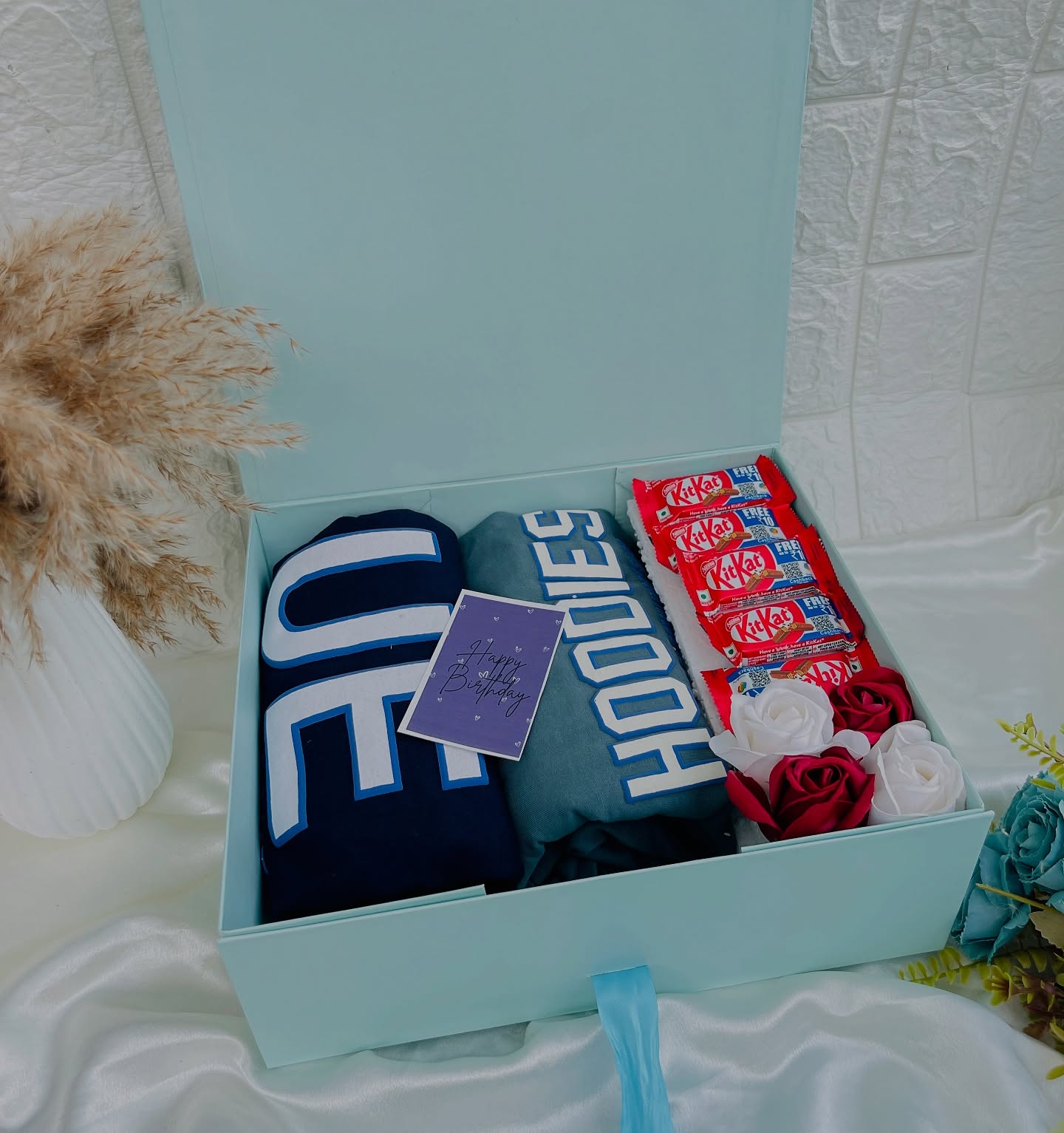 A mint-blue gift box with two cozy statement hoodies, a stack of KitKat bars, and a Happy Birthday card.